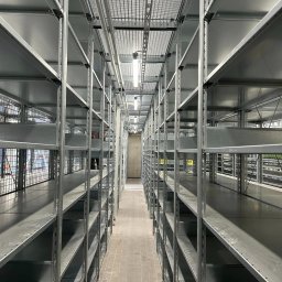 SmartConnectSystems Paweł Wierzelewski - Długi korytarz z rzędami pustych, metalowych regałów magazynowych, oświetlony lampami sufitowymi, z betonową podłogą i widoczną kratką wentylacyjną pod sufitem.