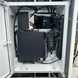 Wnętrze szafki elektrycznej z zainstalowanym routerem TP-Link, rejestratorem wideo i listwą zasilającą, widoczne kable i zasilacze, lekko otwarte białe drzwi szafki.