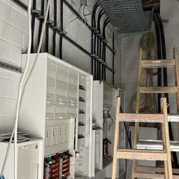 SmartConnectSystems Paweł Wierzelewski - Białe szafy sterownicze z otwartymi drzwiczkami na ścianie z cegły, widoczne kable w korytkach i na drabinkach instalacyjnych oraz drewniane schody przystawne.