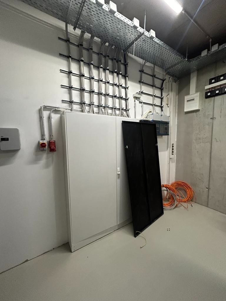 Szafa elektryczna na tle ściany z widocznymi kablami w korytach, pomarańczowe przewody na podłodze.