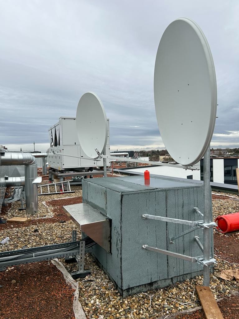 Dwie anteny satelitarne na dachu budynku, widoczne elementy konstrukcyjne i instalacyjne, w tle inne urządzenia i komin wentylacyjny.