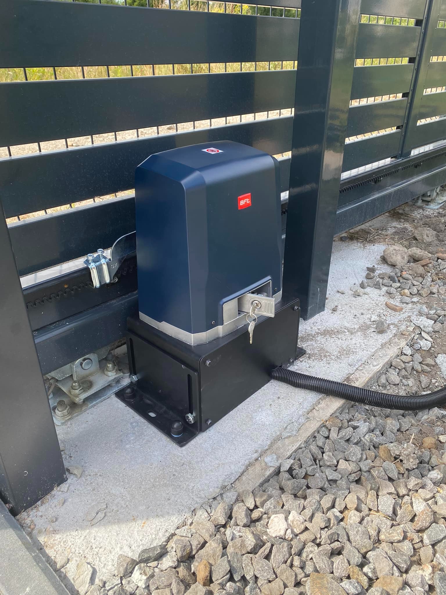 Zamontowany napęd bramy przesuwnej BFT z kluczem w zamku, widoczny przewód zasilający i fragment bramy z metalowymi profilami.