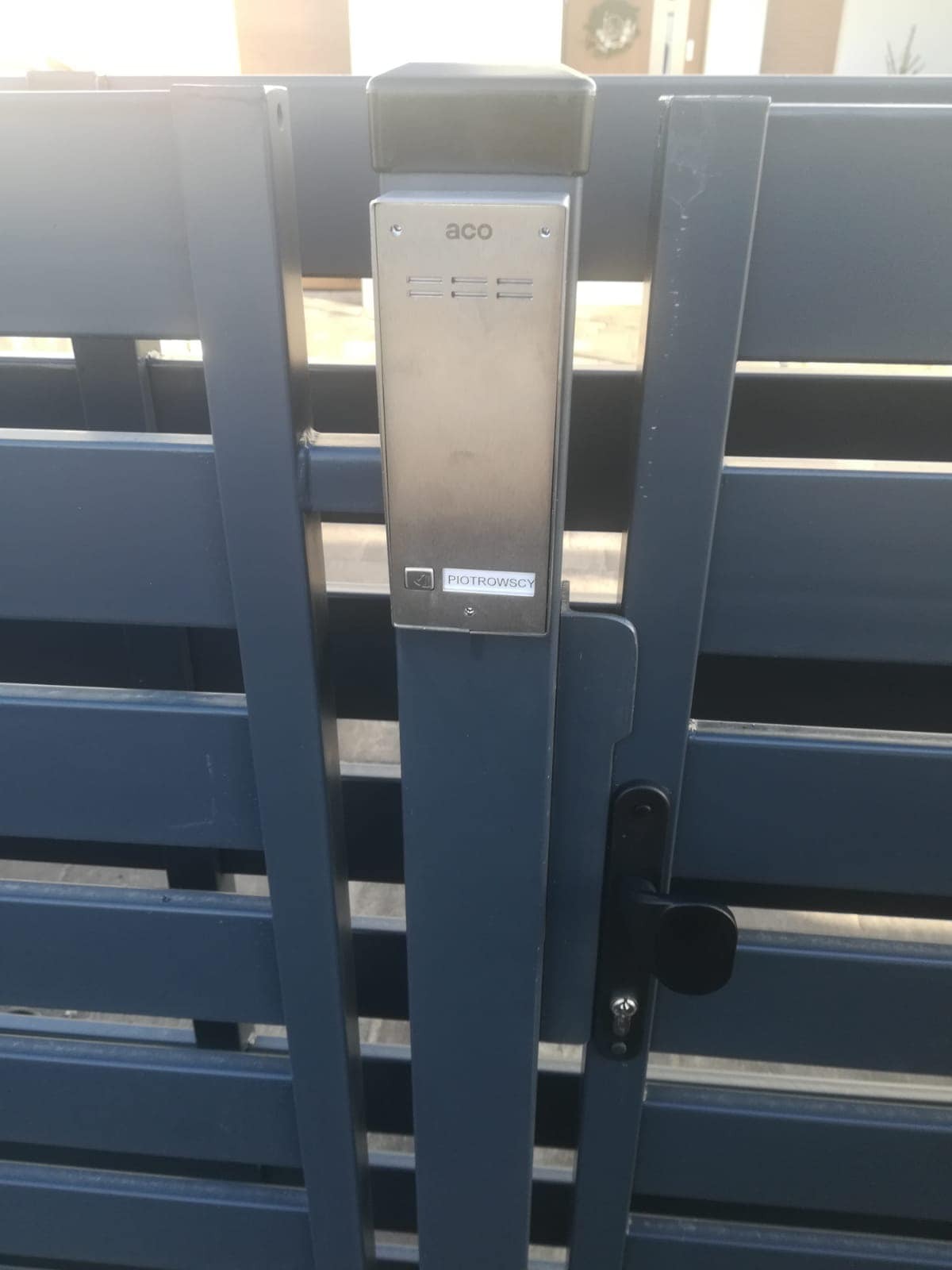 Domofon ACO zamontowany na metalowej, poziomej bramie posesji z czarną klamką. Na domofonie widoczna naklejka z nazwiskiem 'Piotrowscy'.