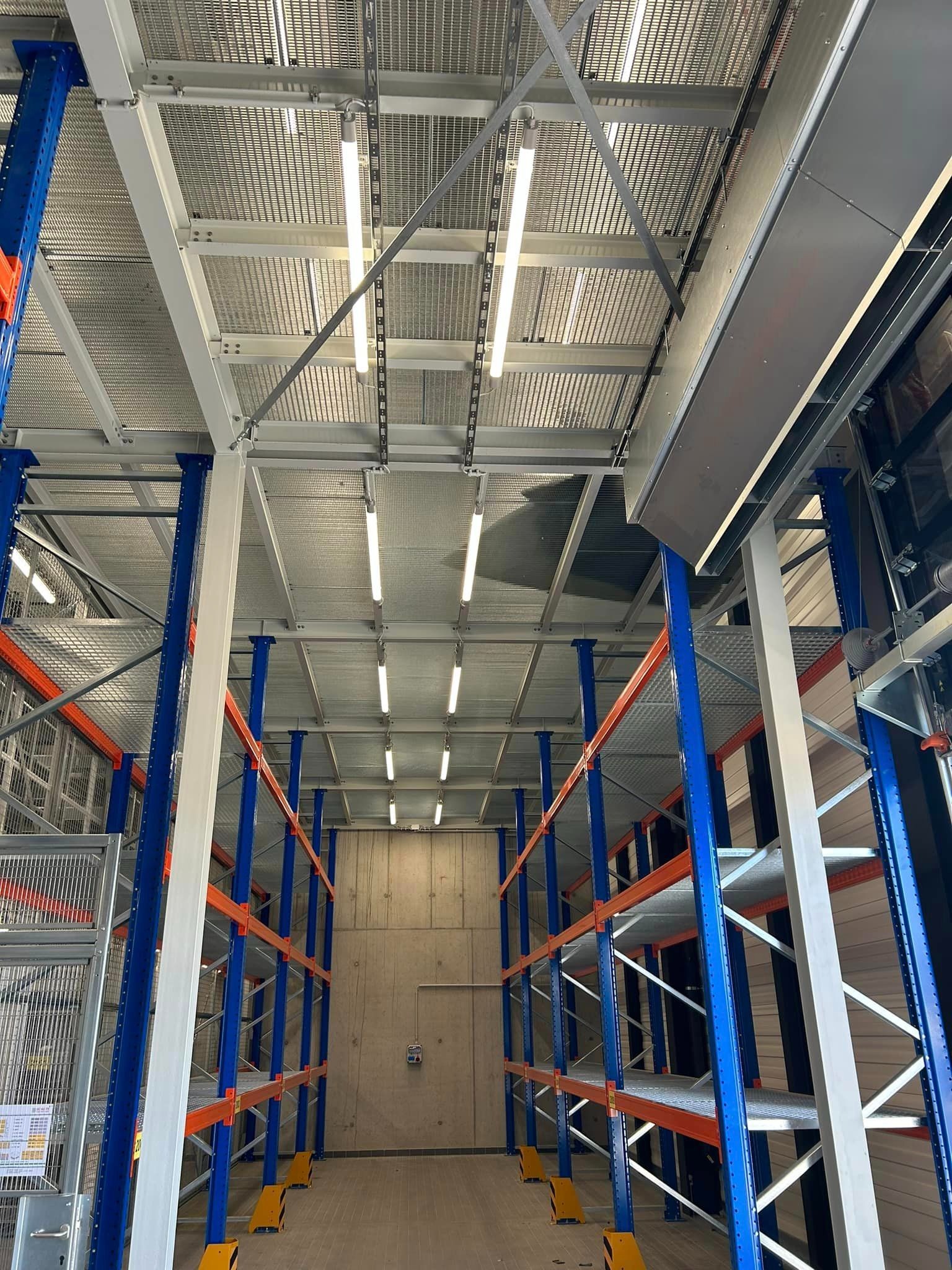Wysokie regały magazynowe z niebieskimi słupkami i pomarańczowymi poprzeczkami, oświetlone lampami jarzeniowymi pod ażurowym sufitem w hali.