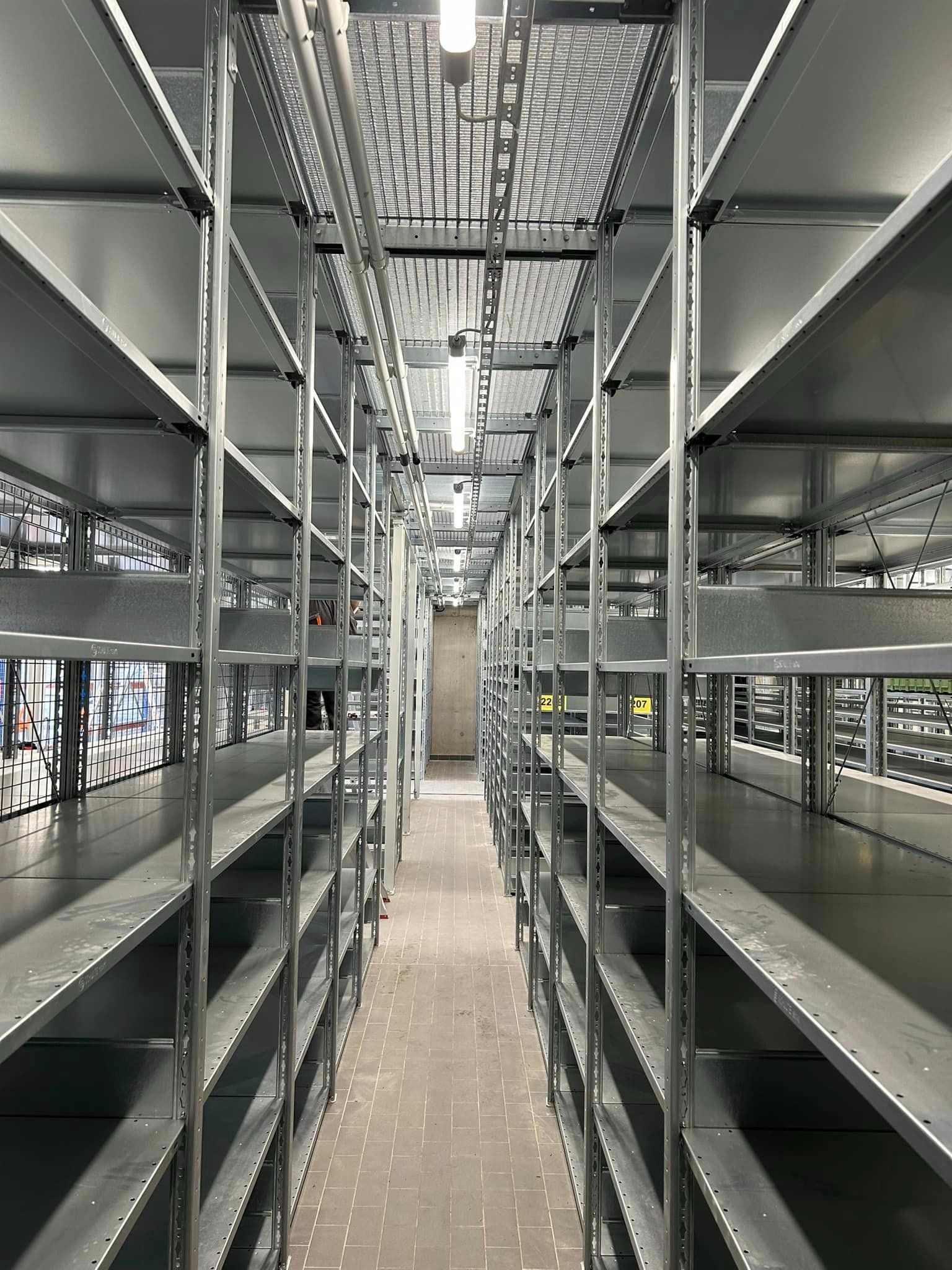 Długi korytarz z rzędami pustych, metalowych regałów magazynowych, oświetlony lampami sufitowymi, z betonową podłogą i widoczną kratką wentylacyjną pod sufitem.