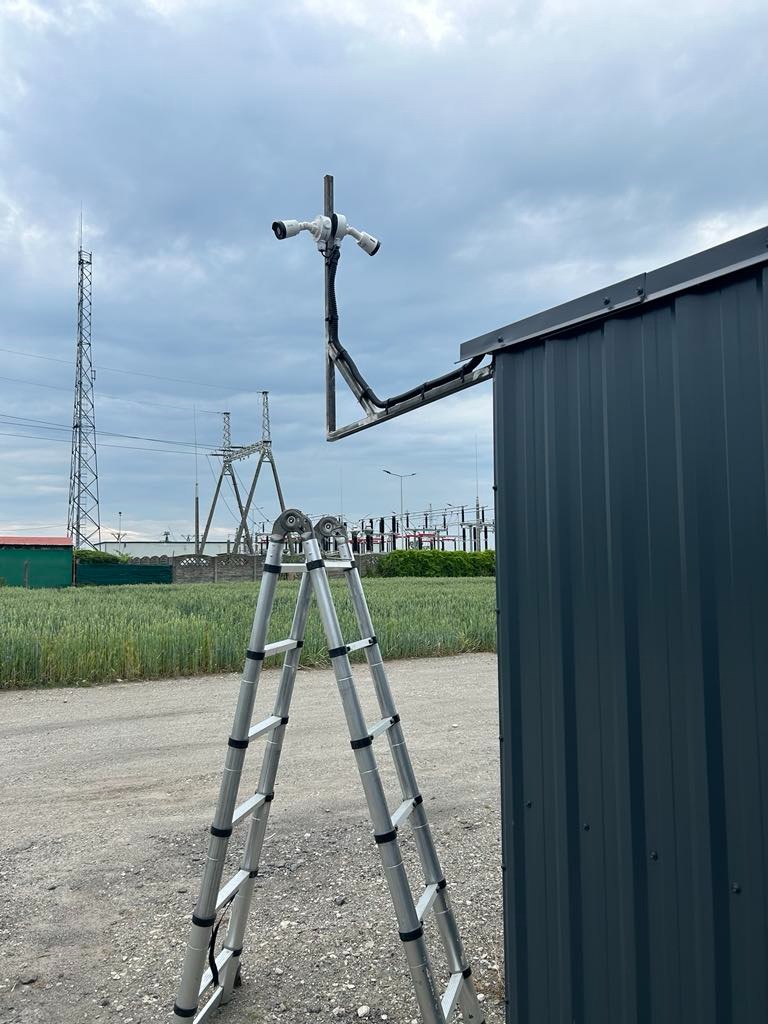 Dwie kamery przemysłowe zamontowane na metalowej konstrukcji przytwierdzonej do boku blaszanej budki, drabina aluminiowa ustawiona obok, w tle pole i słupy wysokiego napięcia.