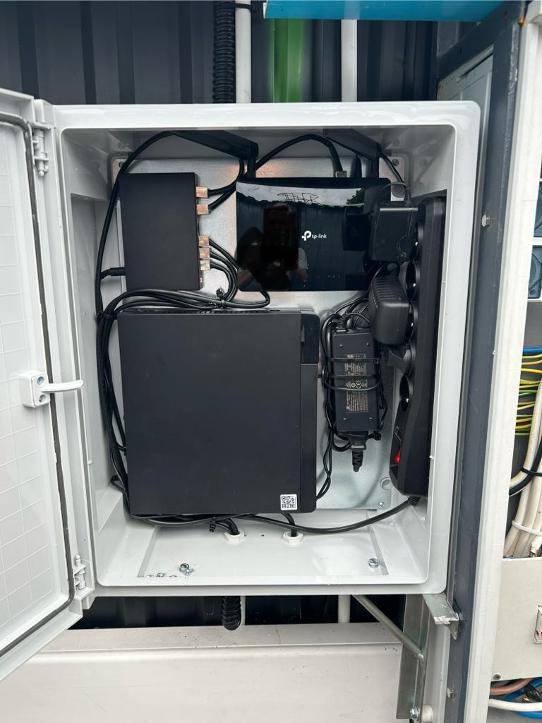 Wnętrze szafki elektrycznej z zainstalowanym routerem TP-Link, rejestratorem wideo i listwą zasilającą, widoczne kable i zasilacze, lekko otwarte białe drzwi szafki.