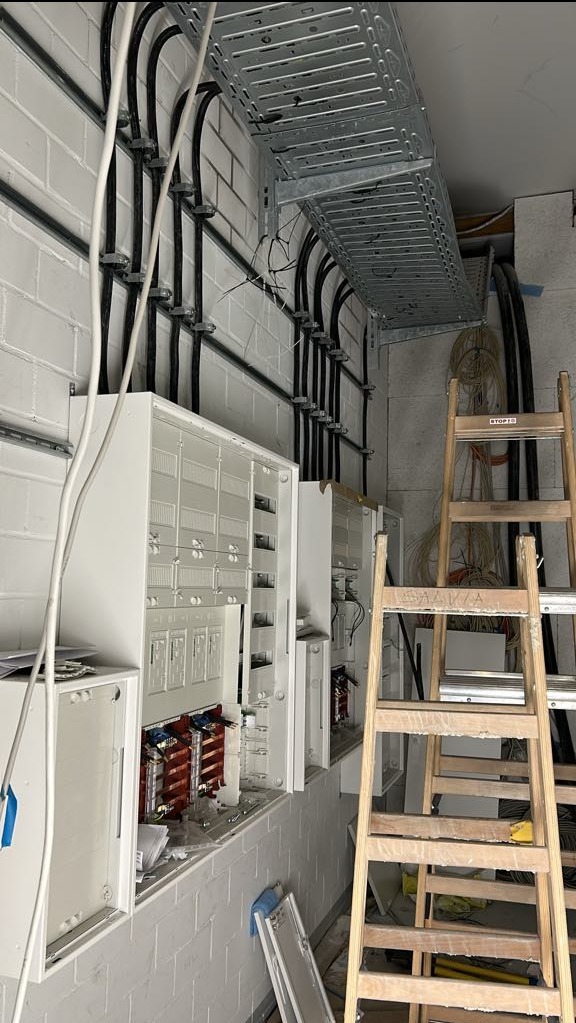 Białe szafy sterownicze z otwartymi drzwiczkami na ścianie z cegły, widoczne kable w korytkach i na drabinkach instalacyjnych oraz drewniane schody przystawne.