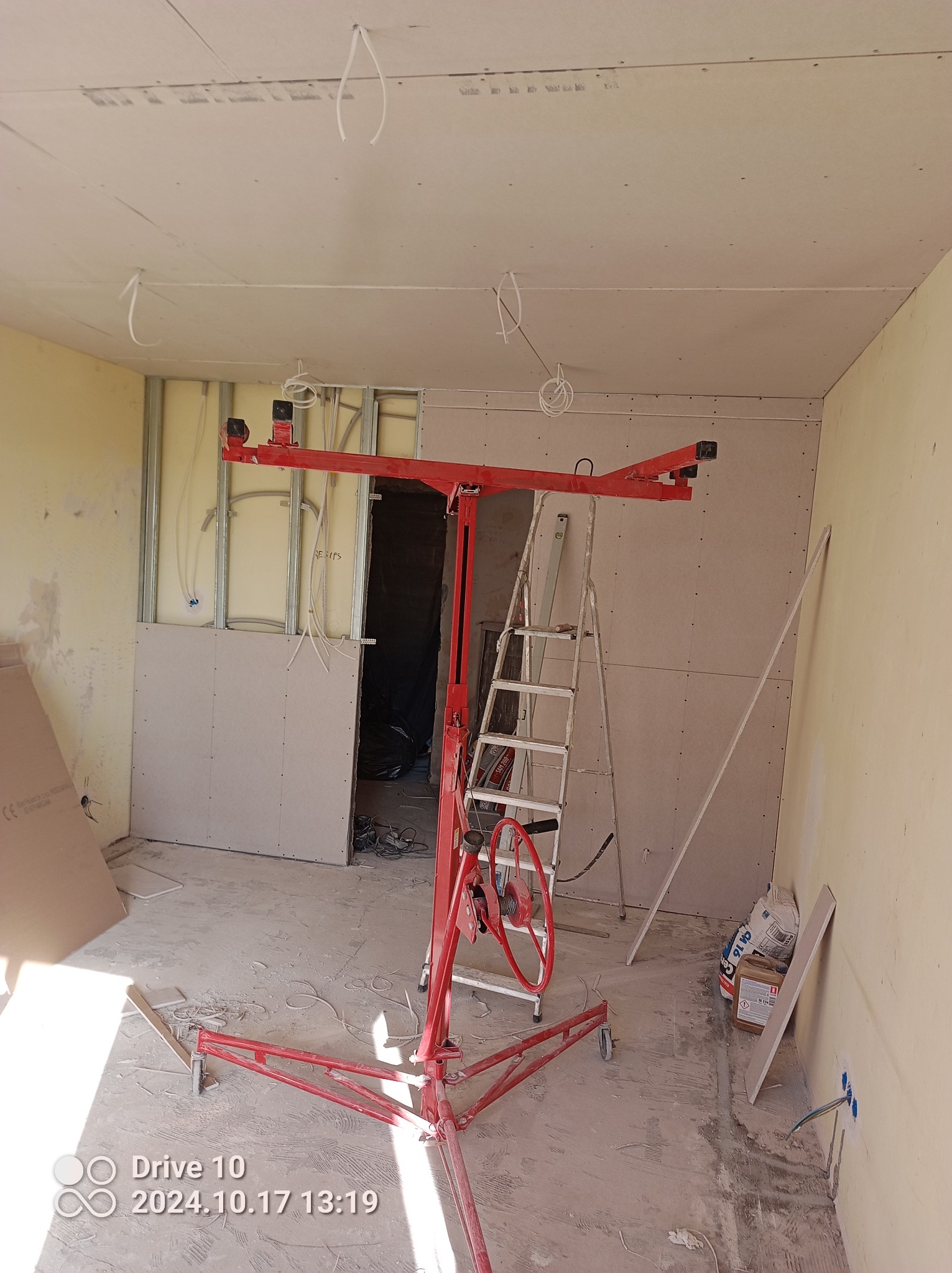Wnętrze w trakcie montażu płyt gipsowo-kartonowych na ścianach i suficie, widoczny podnośnik do płyt w kolorze czerwonym, drabina aluminiowa oraz wystające kable elektryczne.