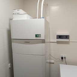 Tomasz Łabędź - Biała pompa ciepła Vaillant w pomieszczeniu z białymi kafelkami, widoczne rury instalacyjne, skrzynka elektryczna i pralka Electrolux.
