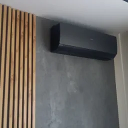 Czarny klimatyzator Haier zamontowany na szarej ścianie z efektem betonu, obok dekoracyjna ścianka z drewnianych listewek, widoczny fragment sufitu z czarnym oświetleniem liniowym.