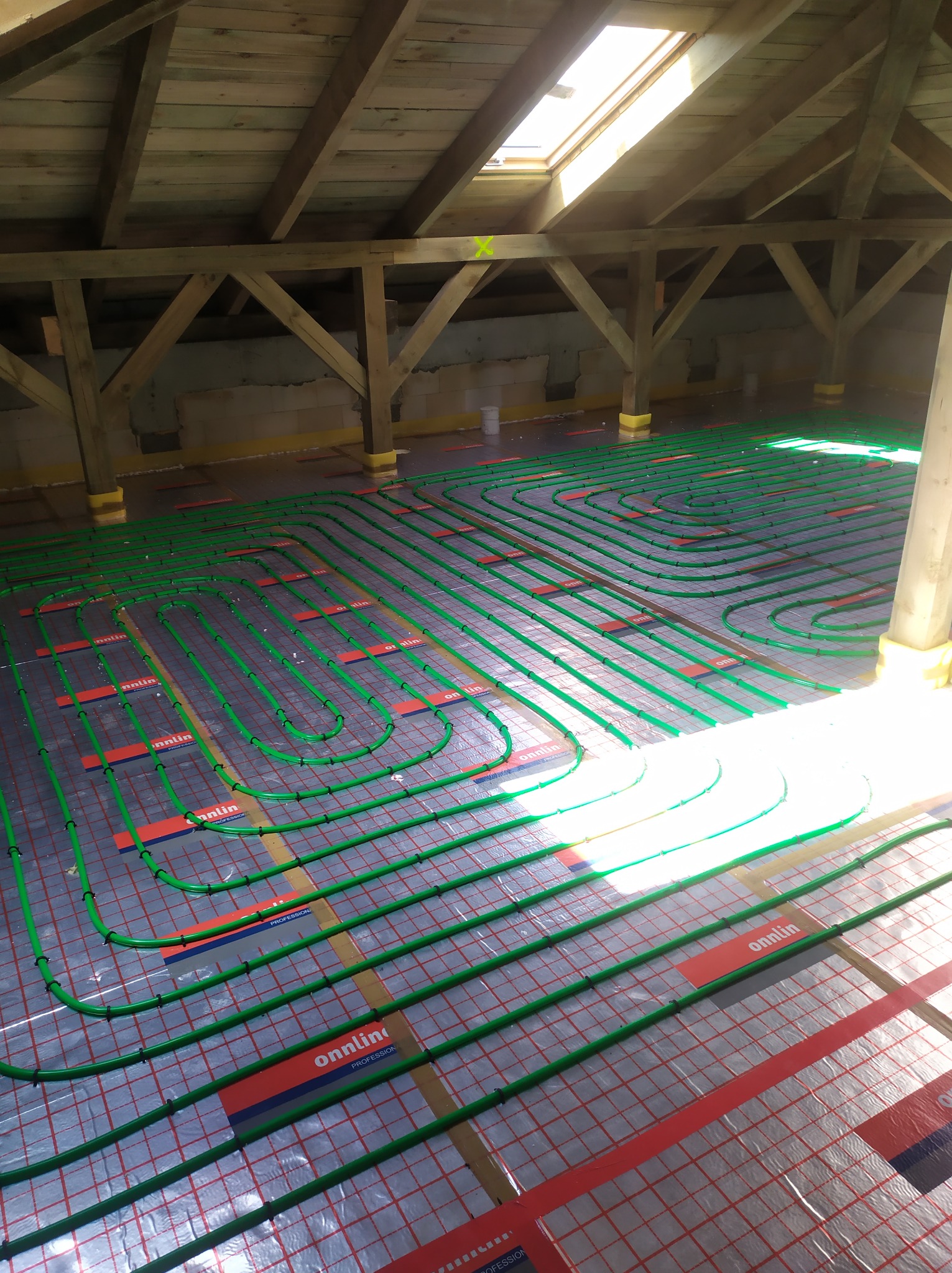 Montaż ogrzewania podłogowego w surowym pomieszczeniu na poddaszu z widoczną konstrukcją drewnianą dachu i świetlikiem.