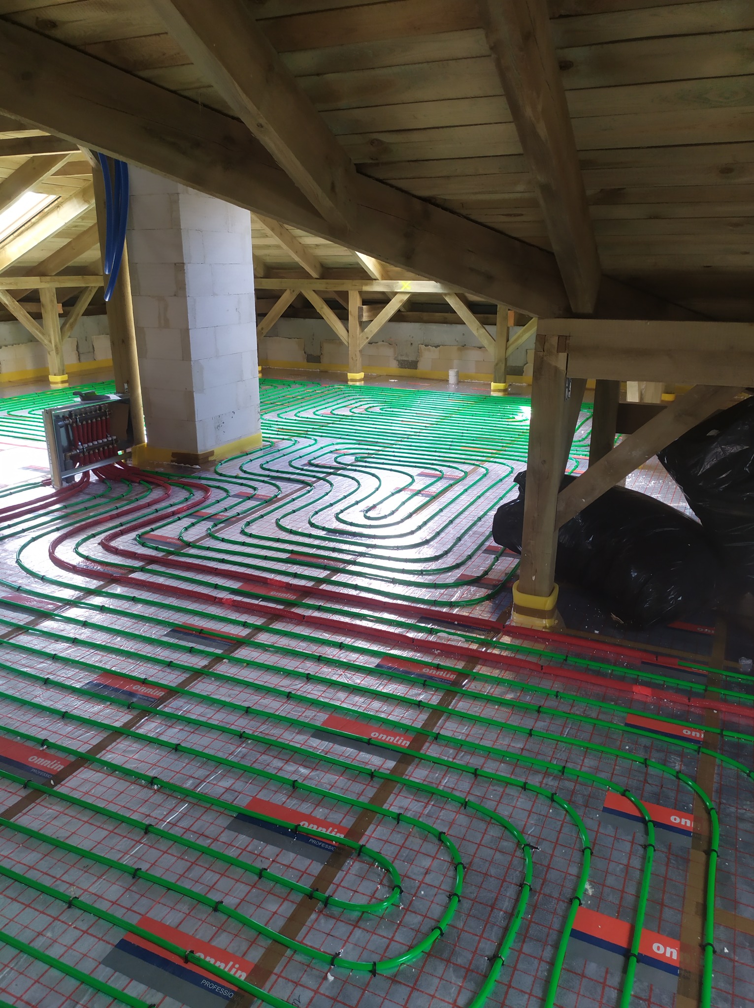 Montaż ogrzewania podłogowego w budynku z drewnianą konstrukcją dachu, widoczne rury grzewcze w kolorze zielonym i czerwonym na folii z nadrukowaną siatką, rozdzielacz w metalowej obudowie przy...