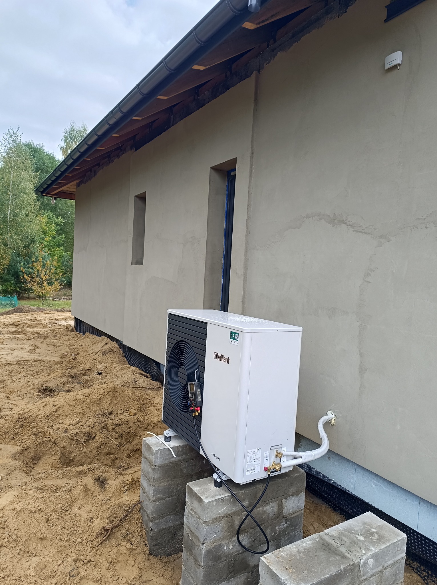 Biała pompa ciepła Vaillant zainstalowana na betonowych bloczkach przy szarej ścianie domu w trakcie budowy, widoczne przyłącza i fragment elewacji.