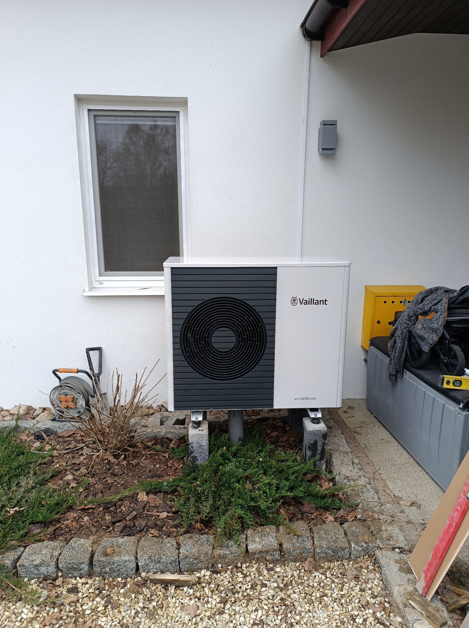 Biała pompa ciepła Vaillant aroTHERM plus zamontowana na betonowych bloczkach przed białą ścianą domu z oknem, obok widoczny żółty skrzynka i narzędzia.