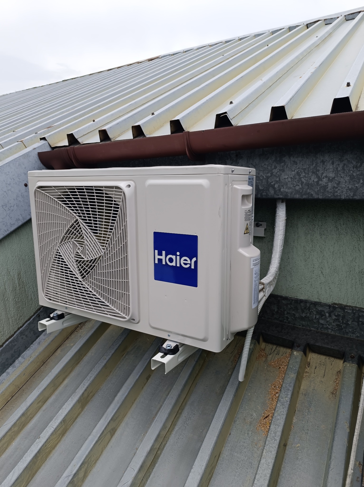 Zewnętrzna jednostka klimatyzacji Haier zamontowana na metalowym dachu z widocznym brązowym orynnowaniem i fragmentami izolacji rur.