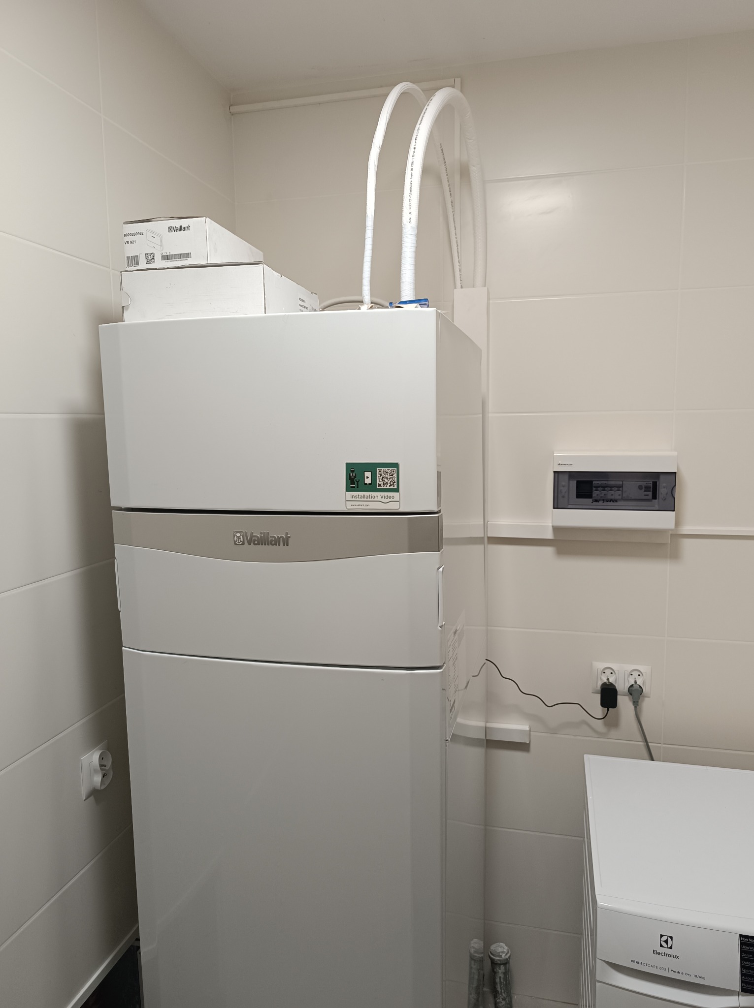 Biała pompa ciepła Vaillant w pomieszczeniu z białymi kafelkami, widoczne rury instalacyjne, skrzynka elektryczna i pralka Electrolux.