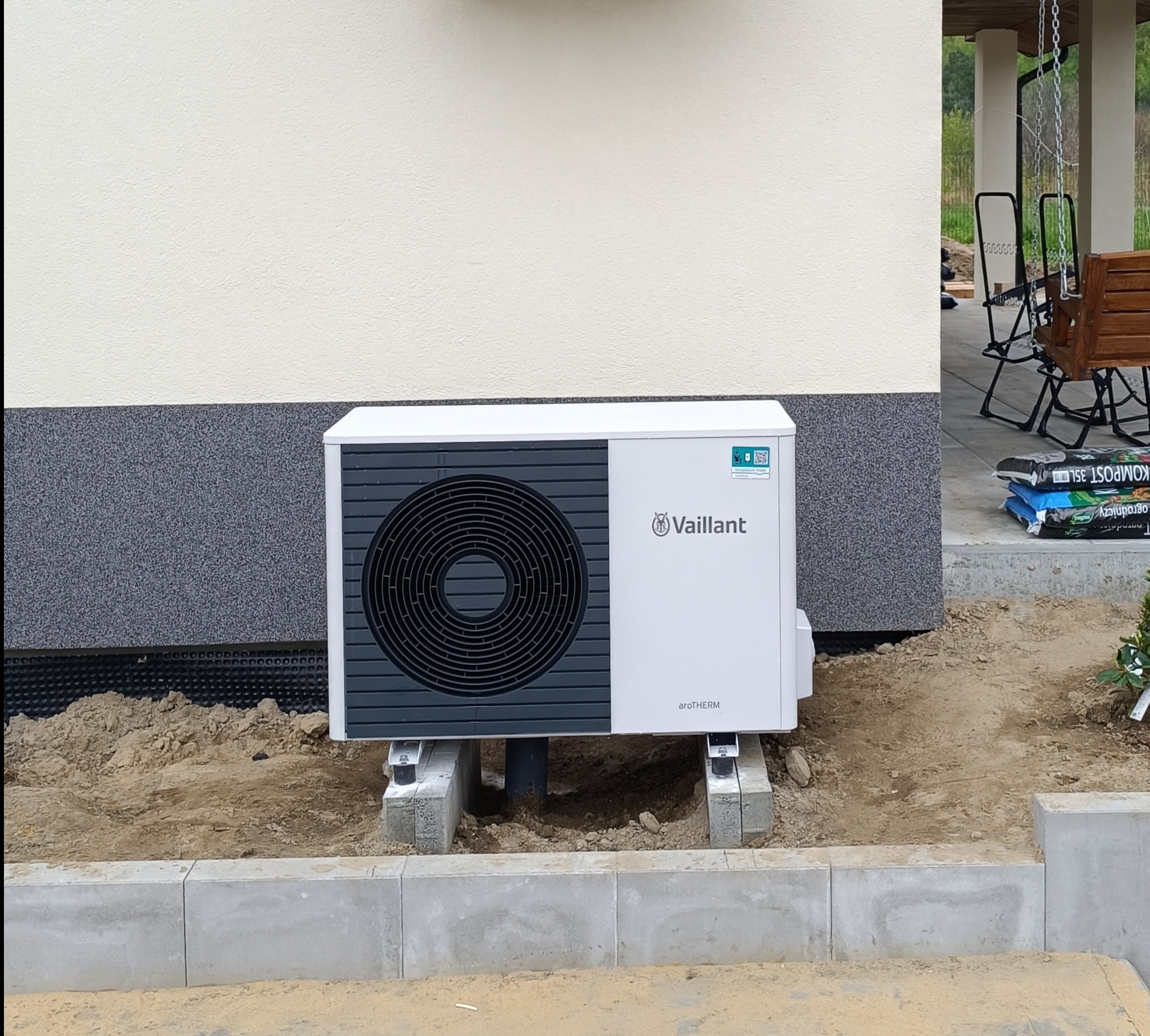 Biała pompa ciepła Vaillant aroTHERM zamontowana na betonowych podstawach przy elewacji domu z widocznym fragmentem ogrodu i workami z kompostem w tle.