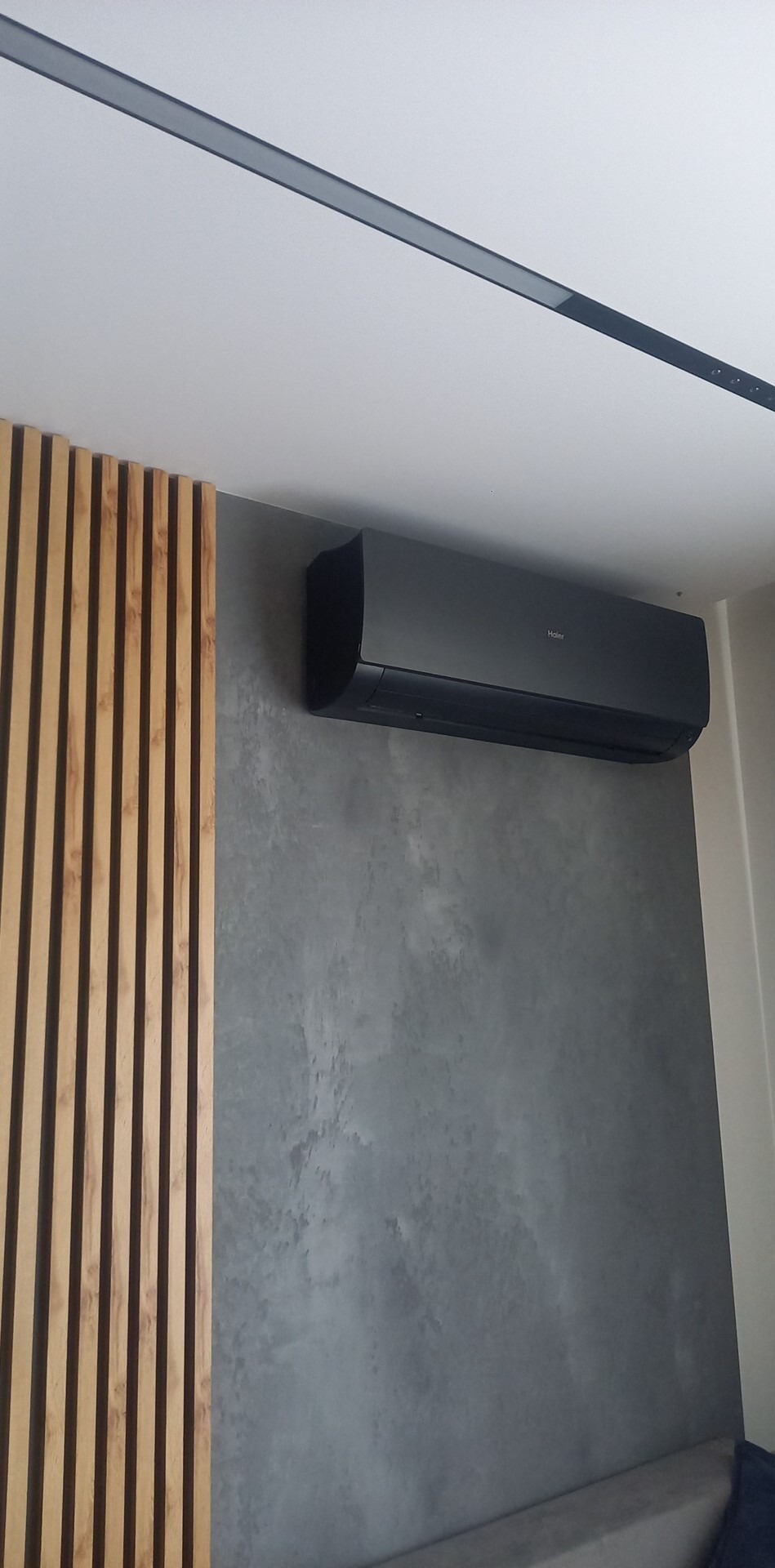 Czarny klimatyzator Haier zamontowany na szarej ścianie z efektem betonu, obok dekoracyjna ścianka z drewnianych listewek, widoczny fragment sufitu z czarnym oświetleniem liniowym.