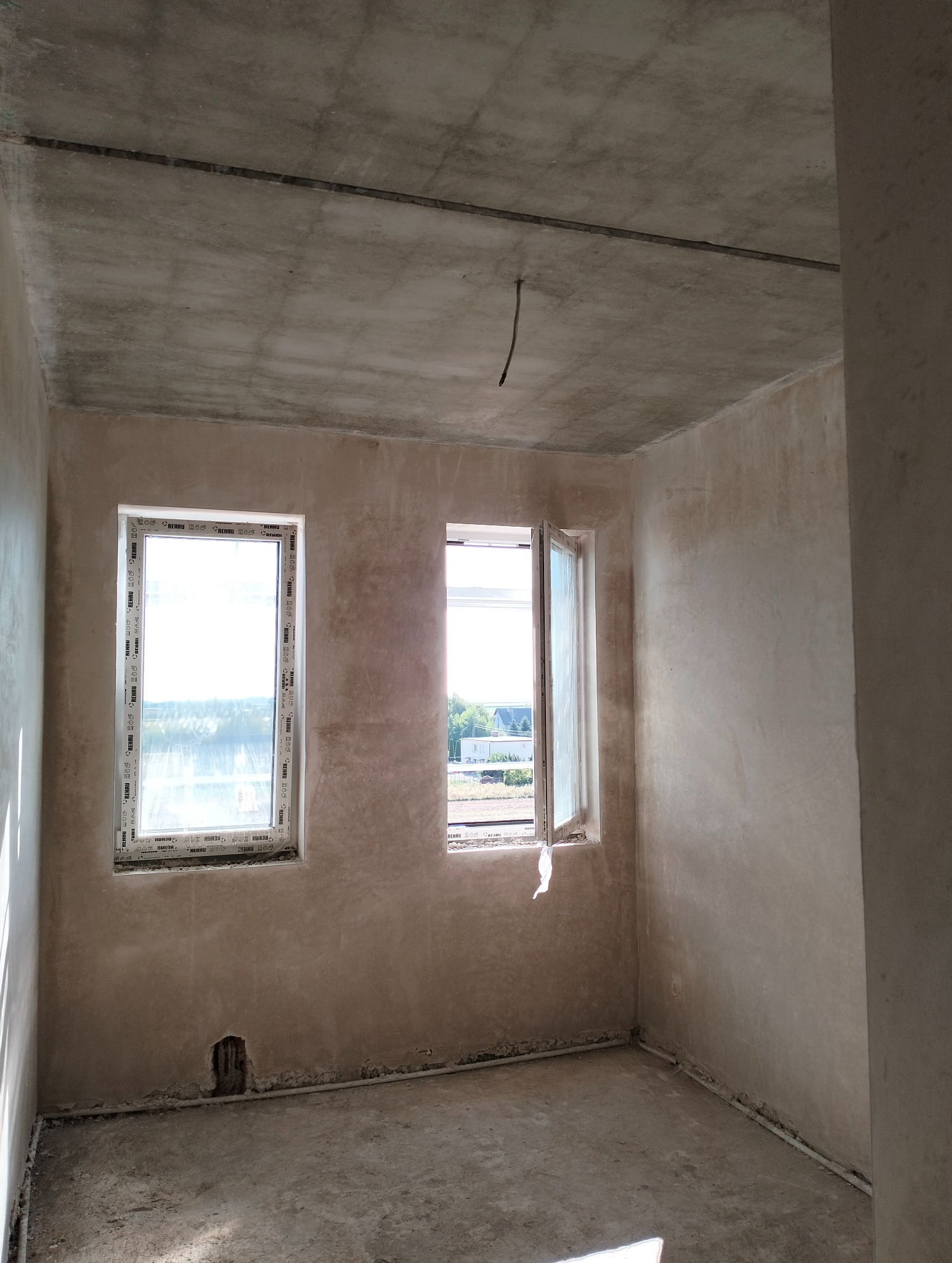 Surowe wnętrze po nałożeniu tynków, z dwoma oknami marki Rehau, jedno z nich uchylone, widoczne instalacje przy podłodze i betonowy sufit z wystającym przewodem.