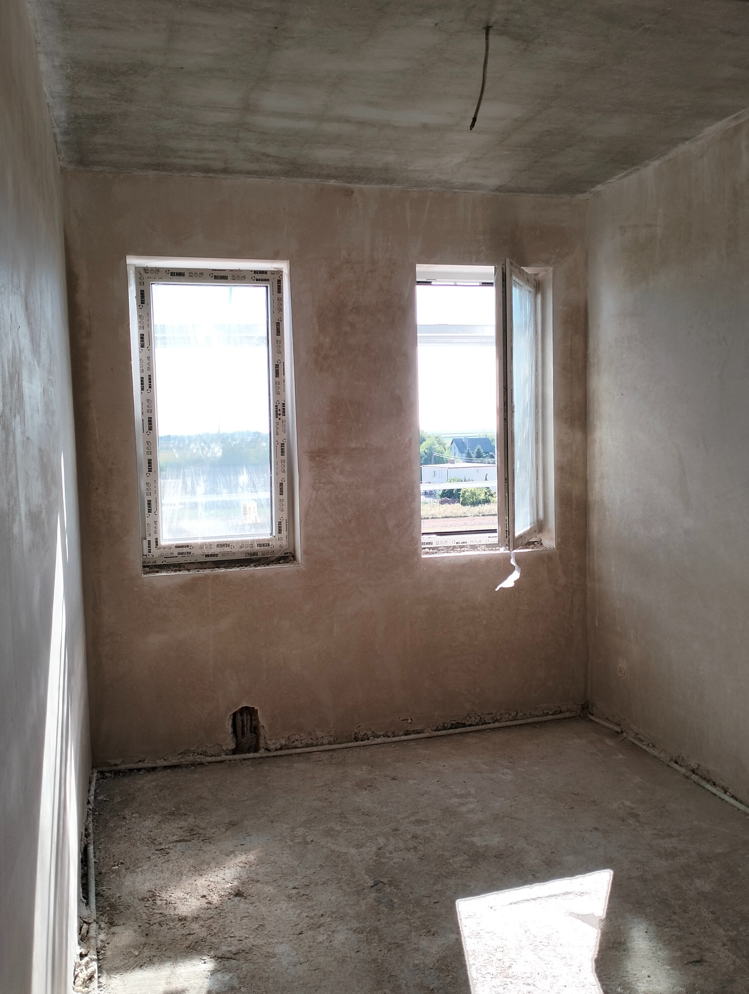 Surowe wnętrze po nałożeniu wstępnej warstwy tynku, widoczne dwa okna z ramami Rehau, odsłonięty kabel zwisający z sufitu i niedoskonałości podłoża.
