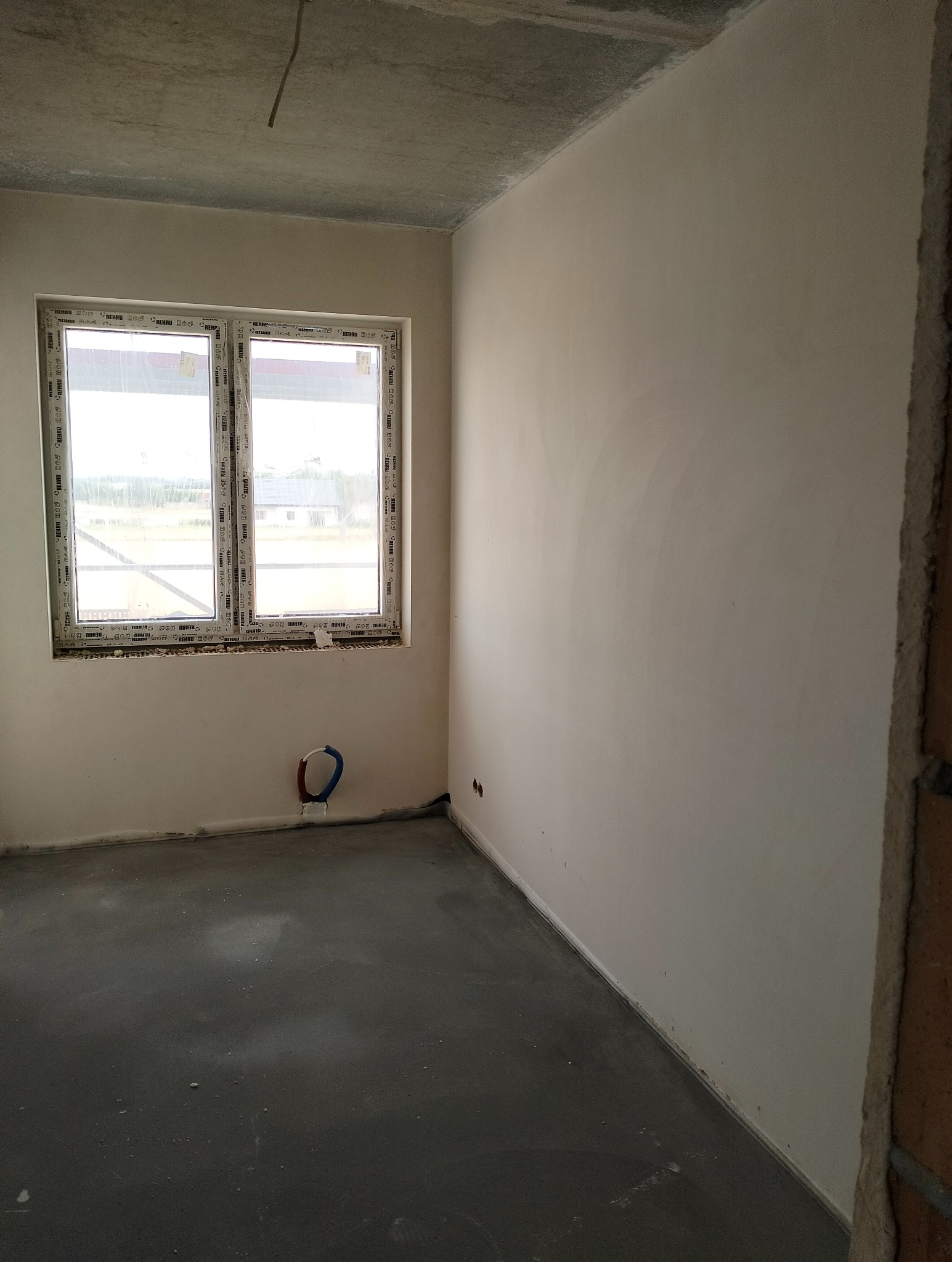 Wnętrze pokoju w stanie deweloperskim po nałożeniu gładzi gipsowej na ściany, widoczne przyłącza hydrauliczne pod oknem i surowy betonowy sufit.