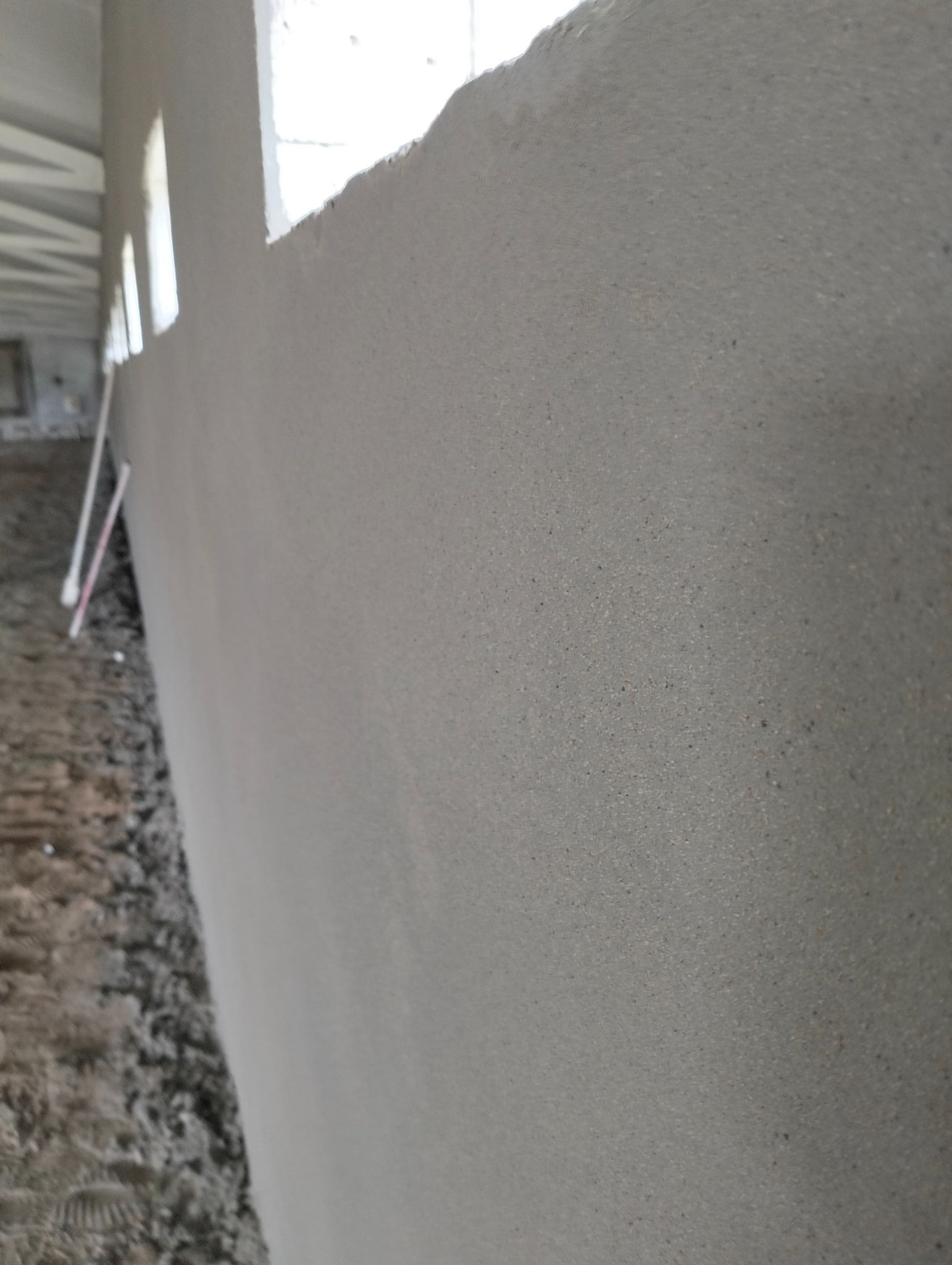 Świeżo nałożony szary tynk na ścianie wewnętrznej, widoczne okno w górnej części kadru, podłoże w stanie surowym.