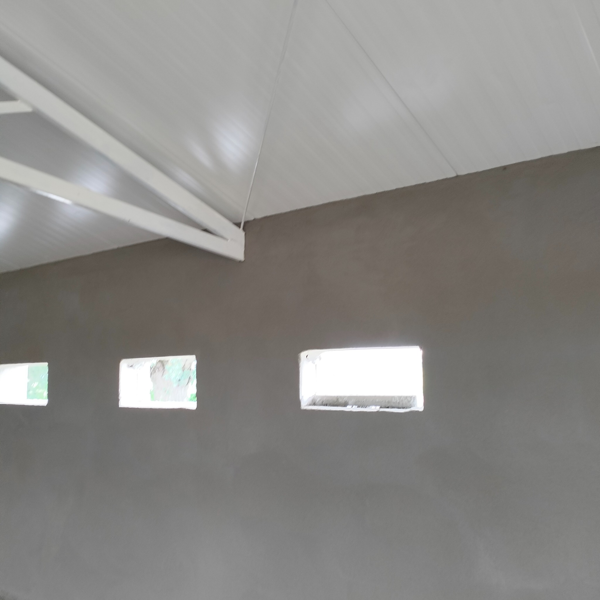 Świeżo otynkowana ściana wewnętrzna z trzema małymi, prostokątnymi otworami okiennymi, widoczne białe elementy konstrukcji dachu.