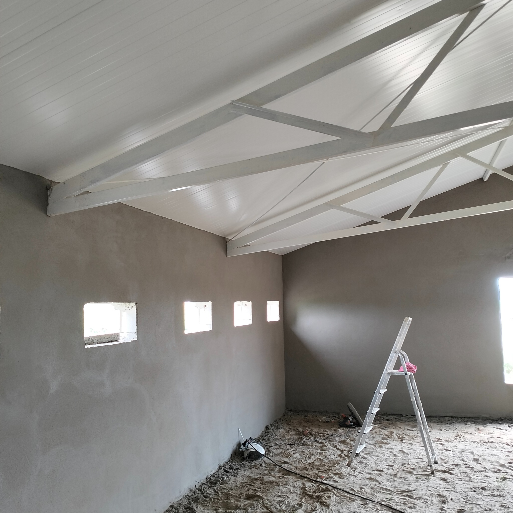 Wnętrze pomieszczenia w trakcie prac wykończeniowych, ściany pokryte szarym tynkiem, sufit z białymi panelami i elementami konstrukcyjnymi, aluminiowa drabina oparta o ścianę, widoczne otwory...