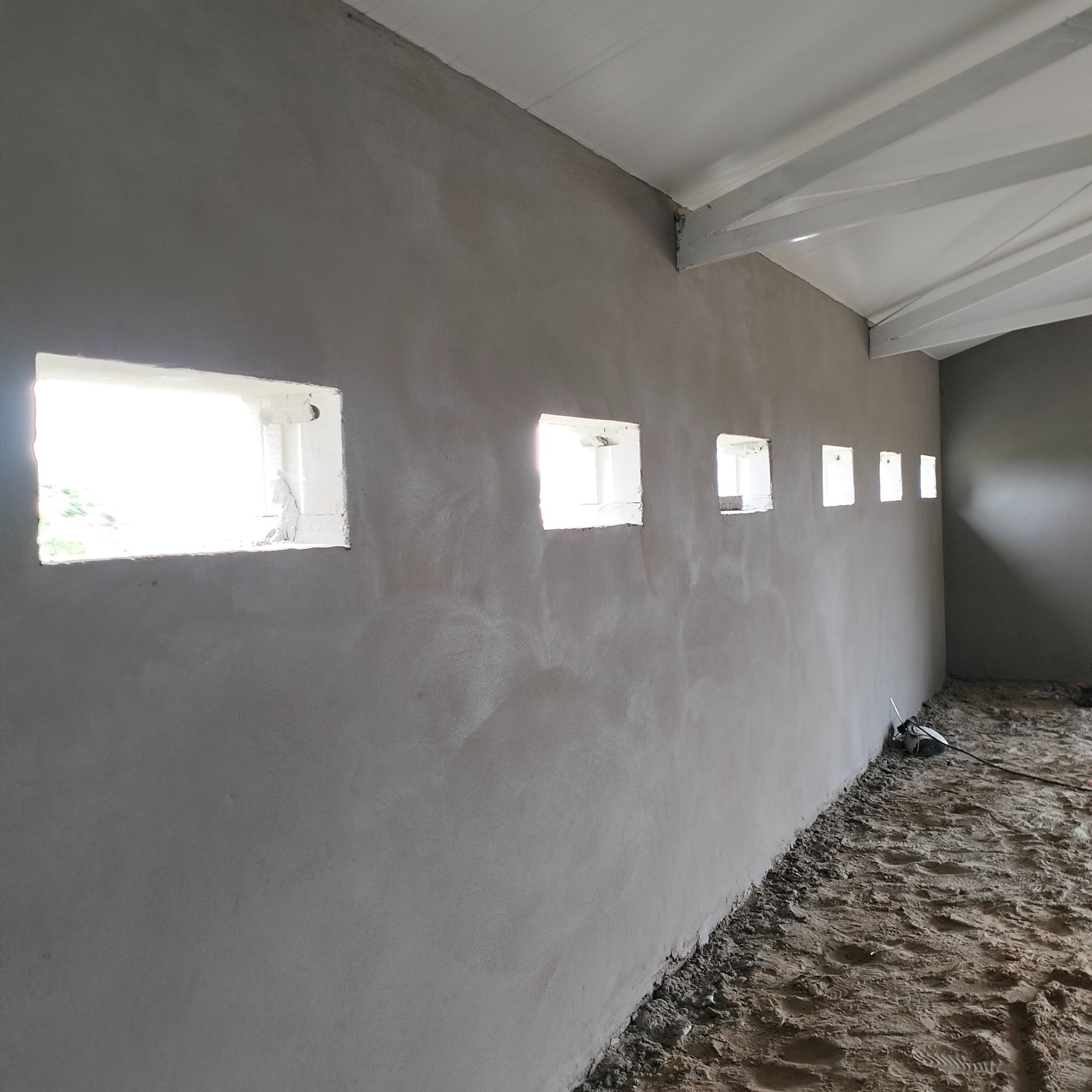Świeżo położony, szary tynk na ścianie wewnętrznej z rzędem małych, kwadratowych okien; widoczne surowe wykończenie wokół okien i nieuprzątnięty gruz na podłodze.
