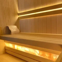 Wnętrze sauny wyłożone jasnym drewnem z podświetleniem solnym pod ławkami i oparciem, oświetlenie LED wzdłuż ściany, wykończenie z płytek imitujących marmur.