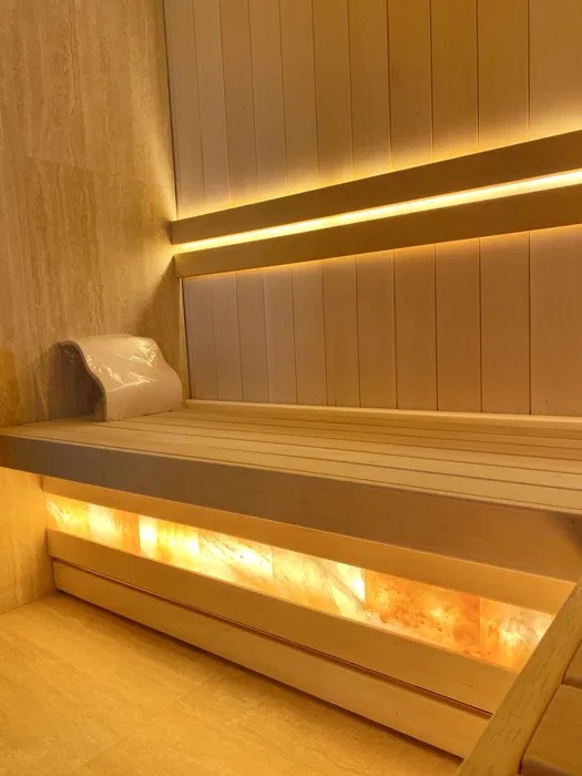 Wnętrze sauny wyłożone jasnym drewnem z podświetleniem solnym pod ławkami i oparciem, oświetlenie LED wzdłuż ściany, wykończenie z płytek imitujących marmur.