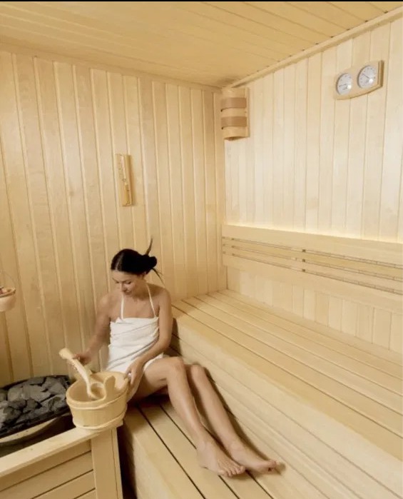 Wnętrze sauny wykończonej jasnym drewnem, z kobietą siedzącą na ławie trzymającą drewniane wiadro i czerpak, widoczne kamienie do polewania, oświetlenie punktowe i zegar z termometrem.