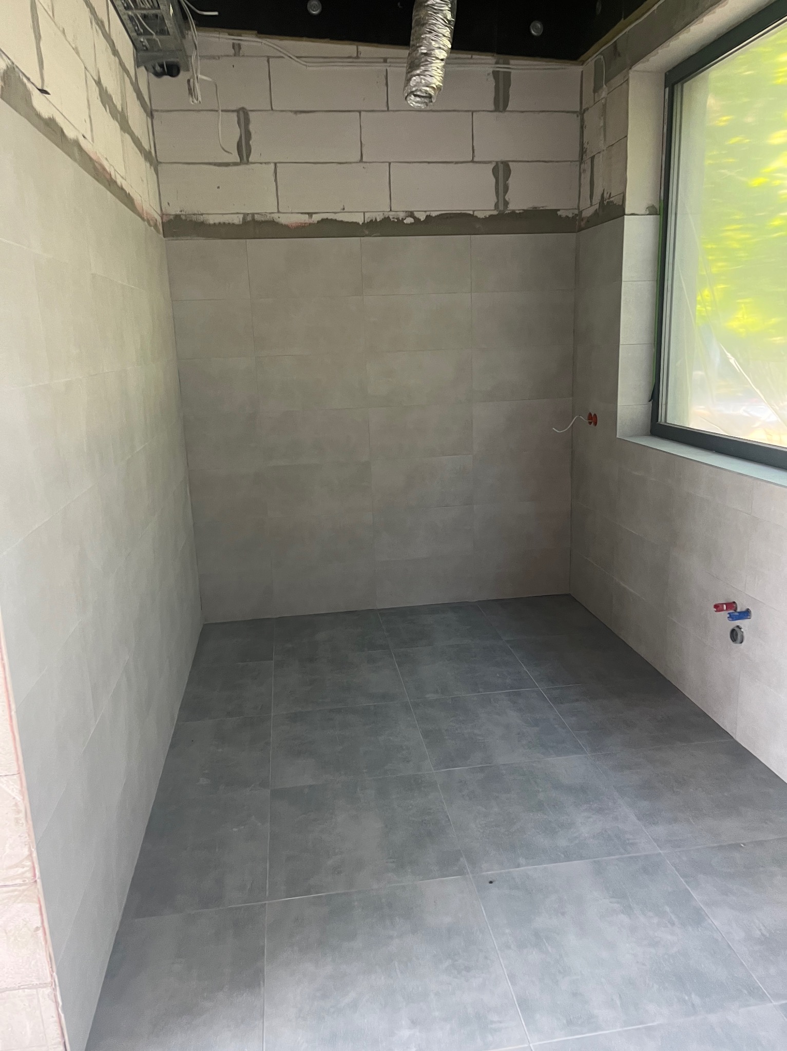 Surowa łazienka w trakcie wykończenia: szare płytki na podłodze i ścianach, widoczne instalacje hydrauliczne i elektryczne, surowy strop z widoczną wentylacją, okno zabezpieczone folią.