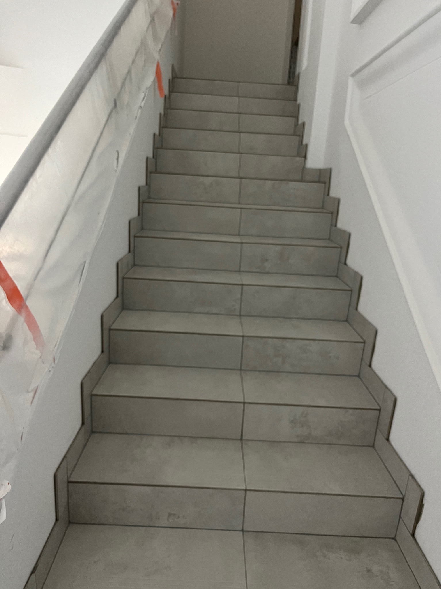 Wnętrze klatki schodowej w trakcie prac wykończeniowych, widoczne schody wyłożone szarą płytką oraz zabezpieczona folią malarską ściana.