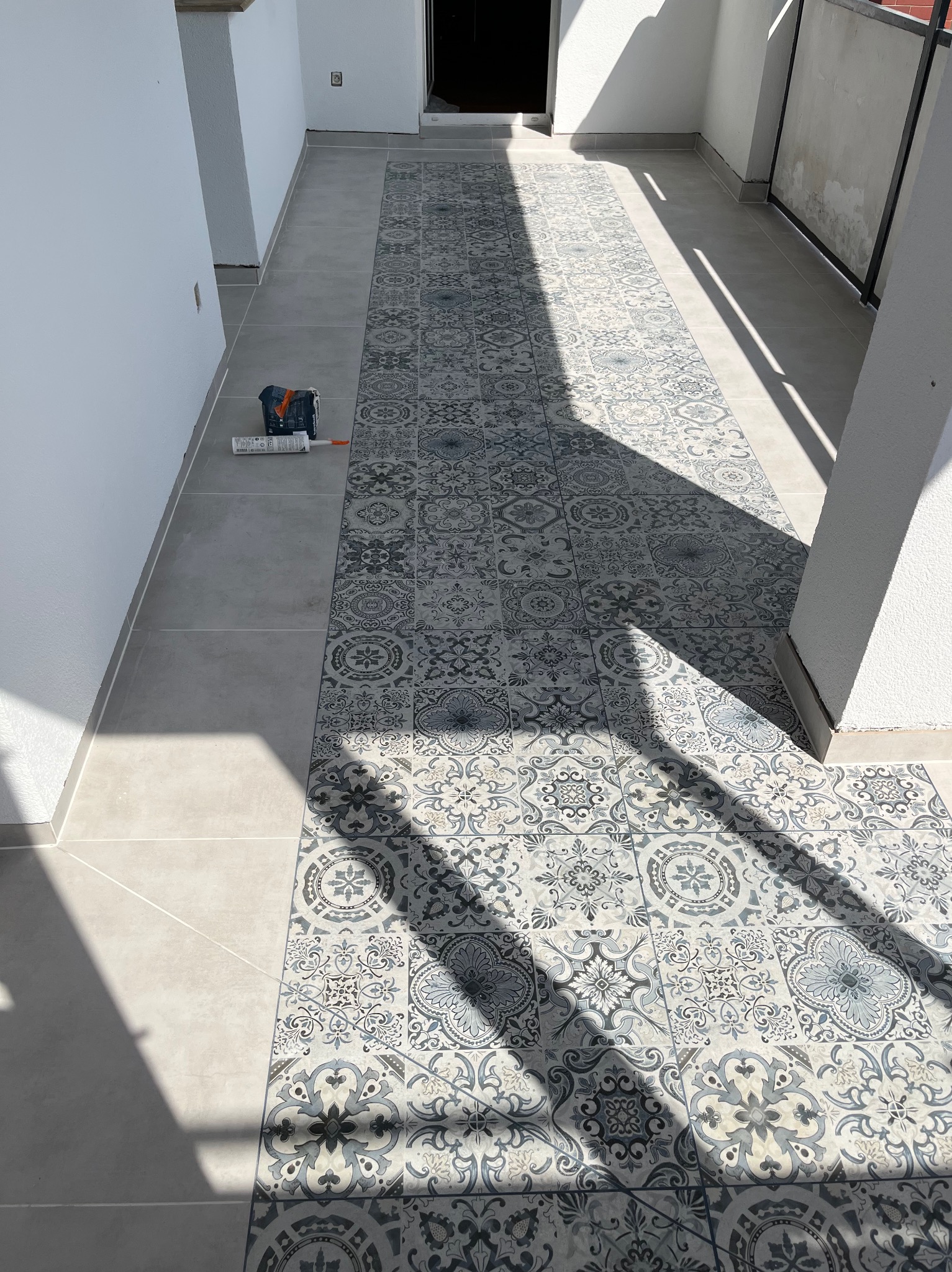 Wykończony balkon z szarymi płytkami w stylu patchwork, widoczne narzędzia do fugowania i cień słoneczny