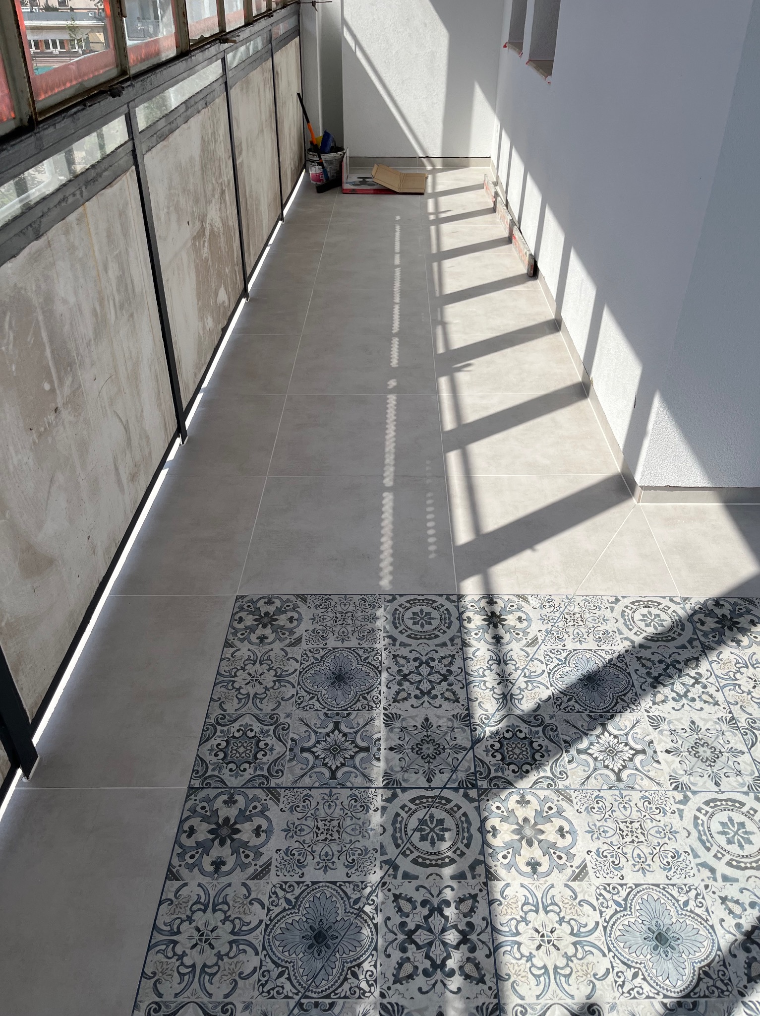 Wyłożony płytkami balkon z wzorzystymi płytkami na podłodze, widoczne narzędzia i materiały budowlane, mocne światło słoneczne tworzy cienie na podłodze.