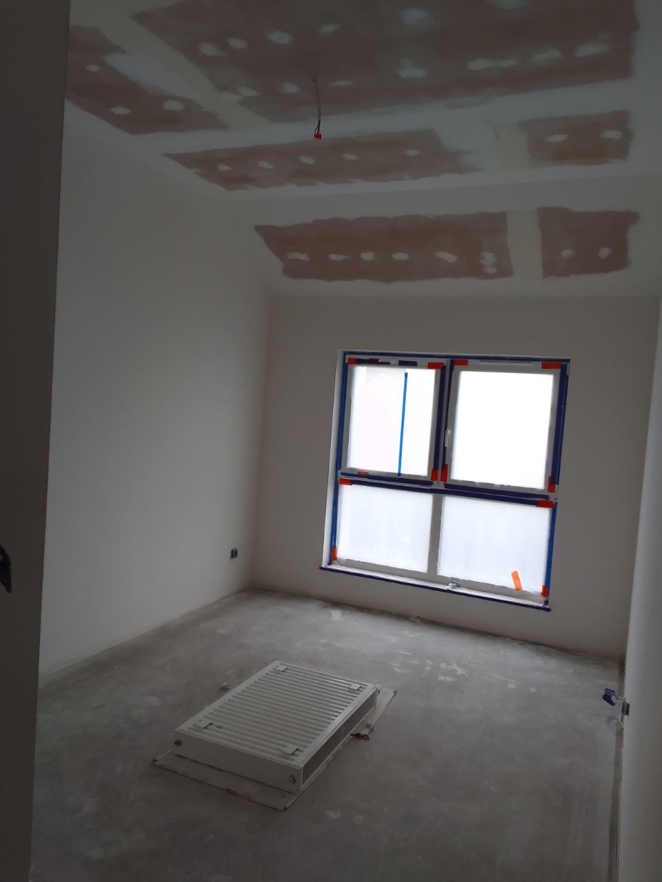 Pokój w trakcie przygotowania pod malowanie: surowy beton na podłodze, grzejnik zabezpieczony kartonem, okno oklejone folią, ślady po łataniu ubytków na suficie.