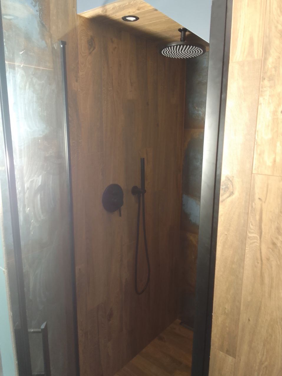 Kabina prysznicowa wykończona panelami imitującymi drewno, z czarną armaturą: deszczownica i bateria z wężem. Widoczne oświetlenie punktowe w suficie i fragment szklanych drzwi kabiny.