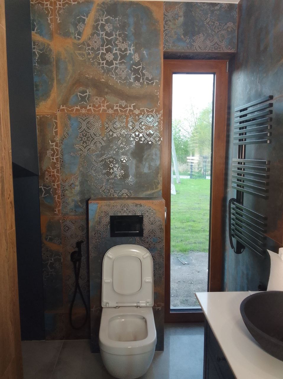 Nowoczesna toaleta podwieszana z otwartą klapą, obudowana płytkami z wzorem w odcieniach niebieskiego i rdzy, na ścianie grzejnik drabinkowy w ciemnym kolorze, widok na zewnątrz przez okno.