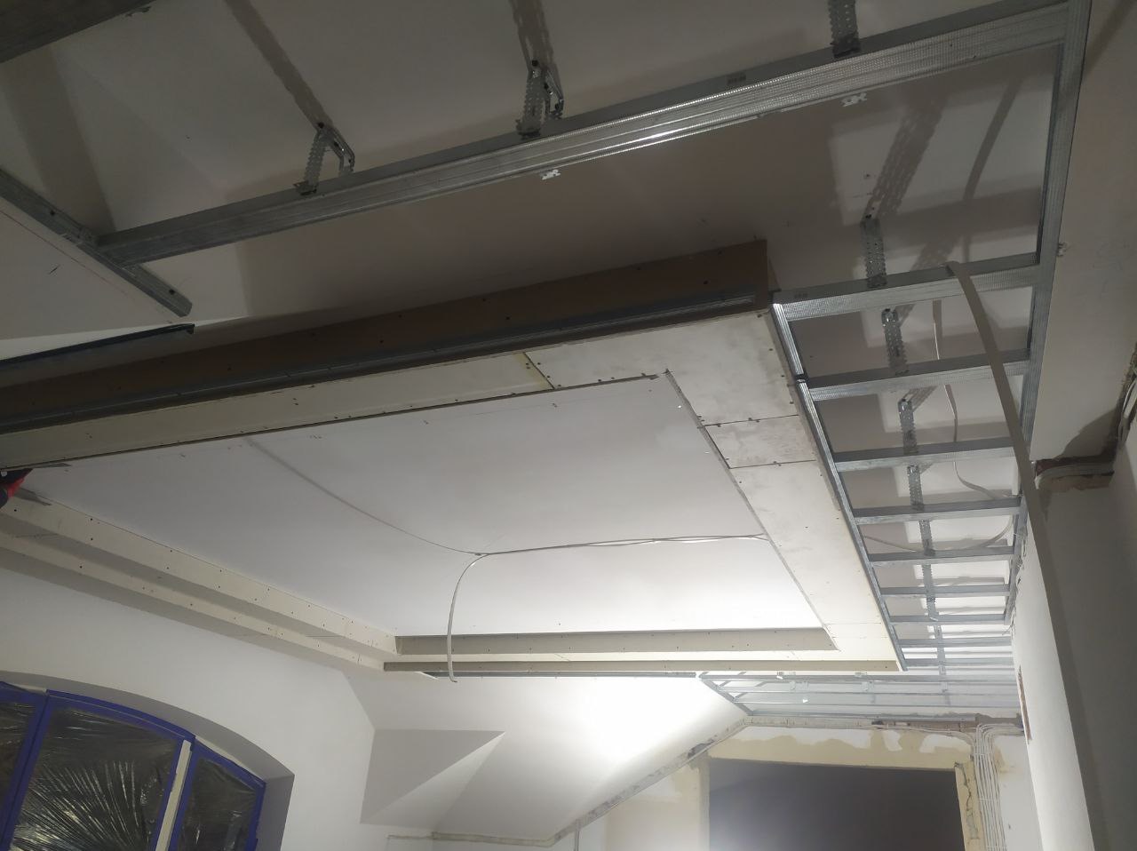 Montaż sufitu podwieszanego z płyt gipsowo-kartonowych na metalowej konstrukcji, widoczne kable elektryczne i częściowo zabudowane oświetlenie.