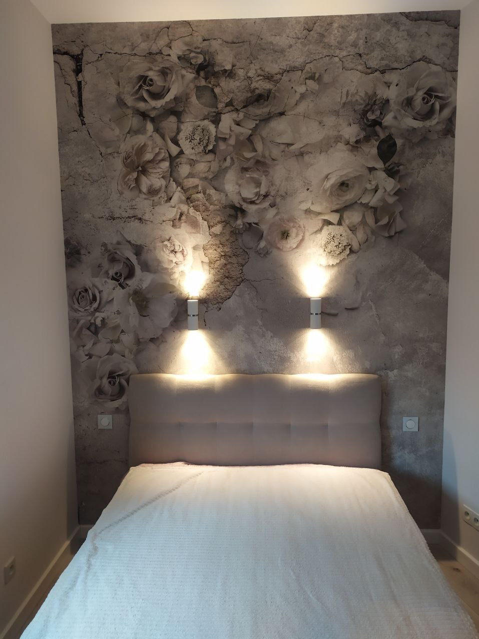 Sypialnia z łóżkiem i tapetą w motywie kwiatowym, oświetlona dwoma kinkietami ściennymi z ciepłym światłem.