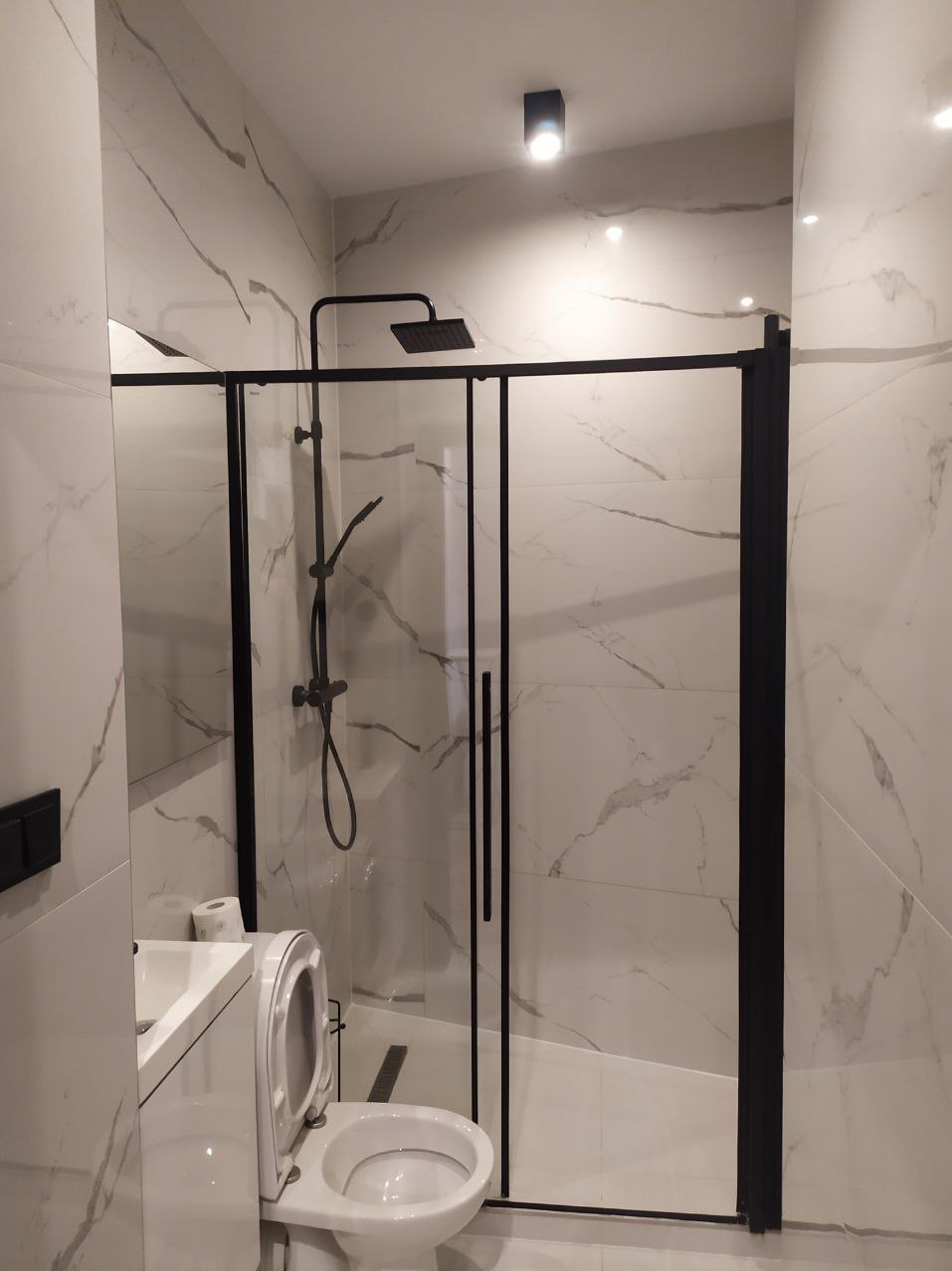 Wykończona łazienka z kabiną prysznicową typu walk-in z czarnymi profilami, białe płytki imitujące marmur, toaleta i umywalka w minimalistycznym stylu.