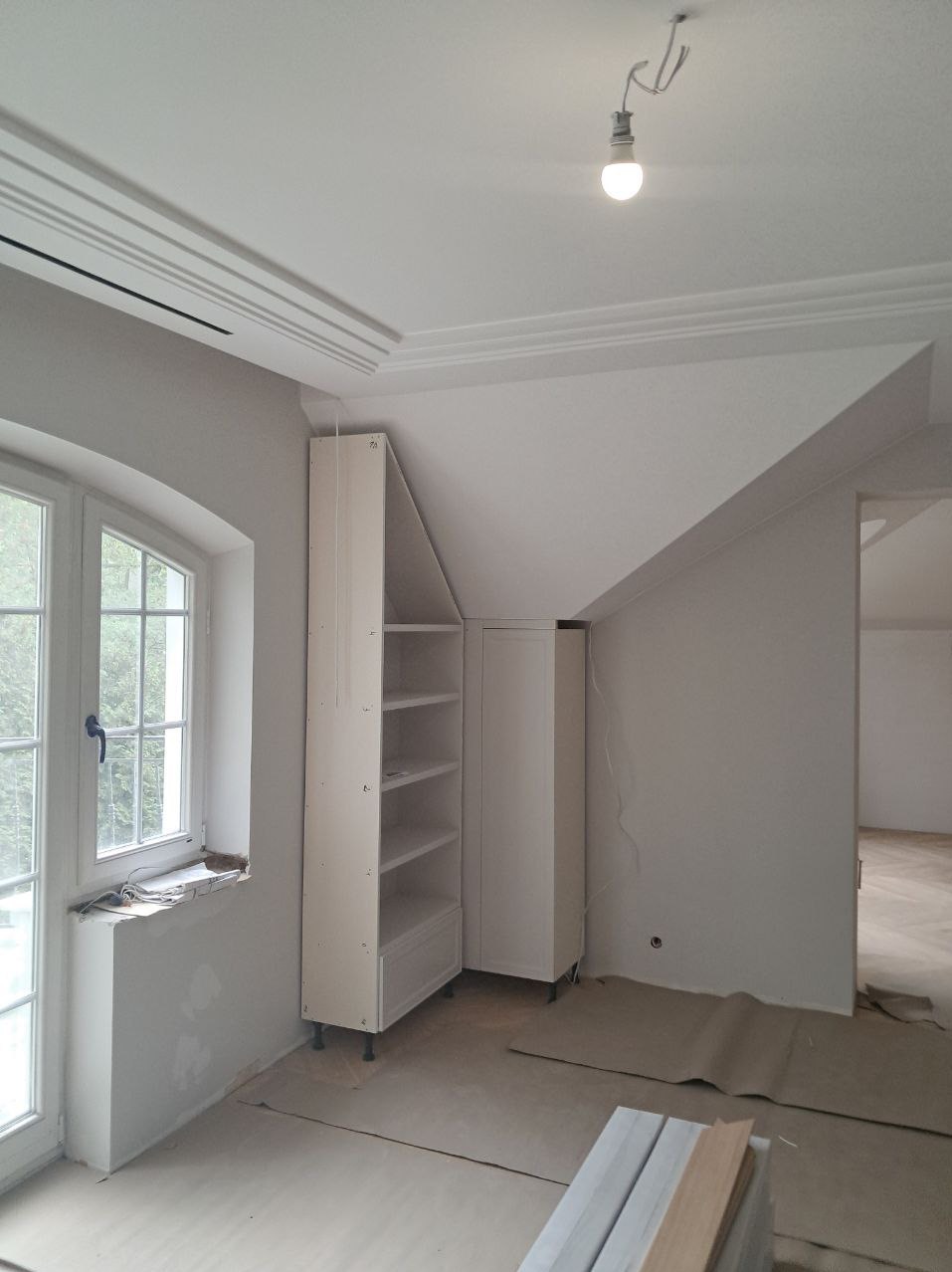 Wnętrze pokoju na poddaszu z oknem, wykończonymi ścianami i sufitem z dekoracyjnymi listwami, z zamontowanymi białymi szafkami i przygotowanymi panelami podłogowymi.