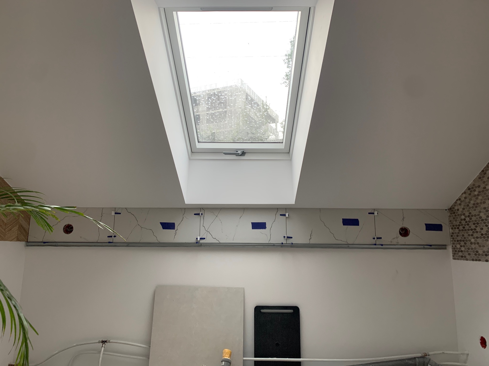 Montaż płytek imitujących marmur na ścianie, widoczne niebieskie taśmy i krzyżyki dystansowe, okno dachowe z widokiem na budynek w tle.