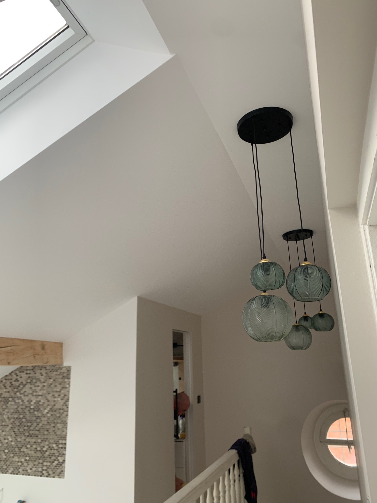 Jasne wnętrze z gładkimi, białymi ścianami, oknem dachowym i wiszącą lampą z kulistymi, żłobionymi kloszami w odcieniach szarości i błękitu.