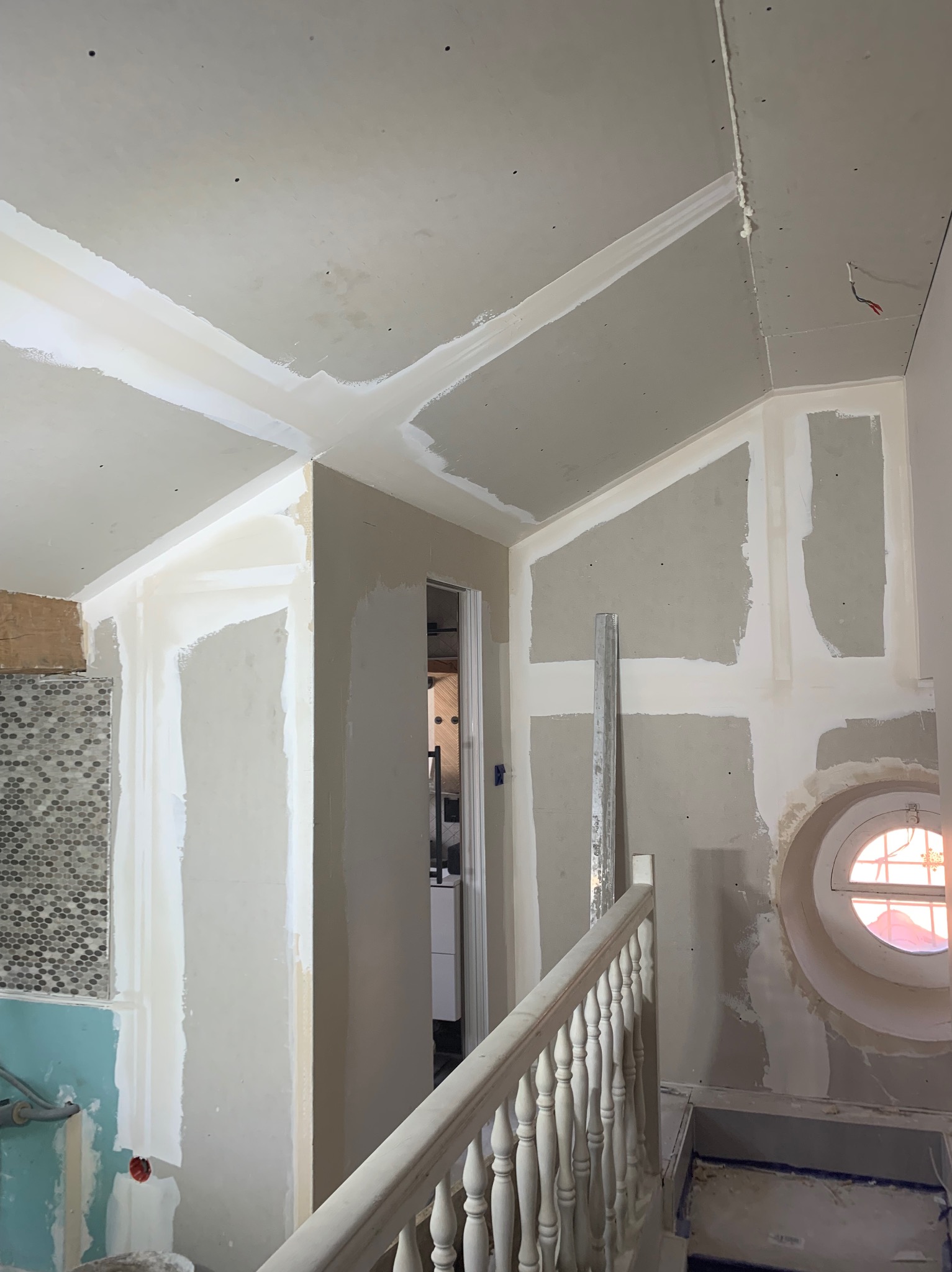 Wnętrze w trakcie prac przygotowawczych do malowania: ściany i sufit z widocznymi łączeniami płyt gipsowo-kartonowych, częściowo pokryte gładzią, z widocznym oknem okrągłym i fragmentem schodów...