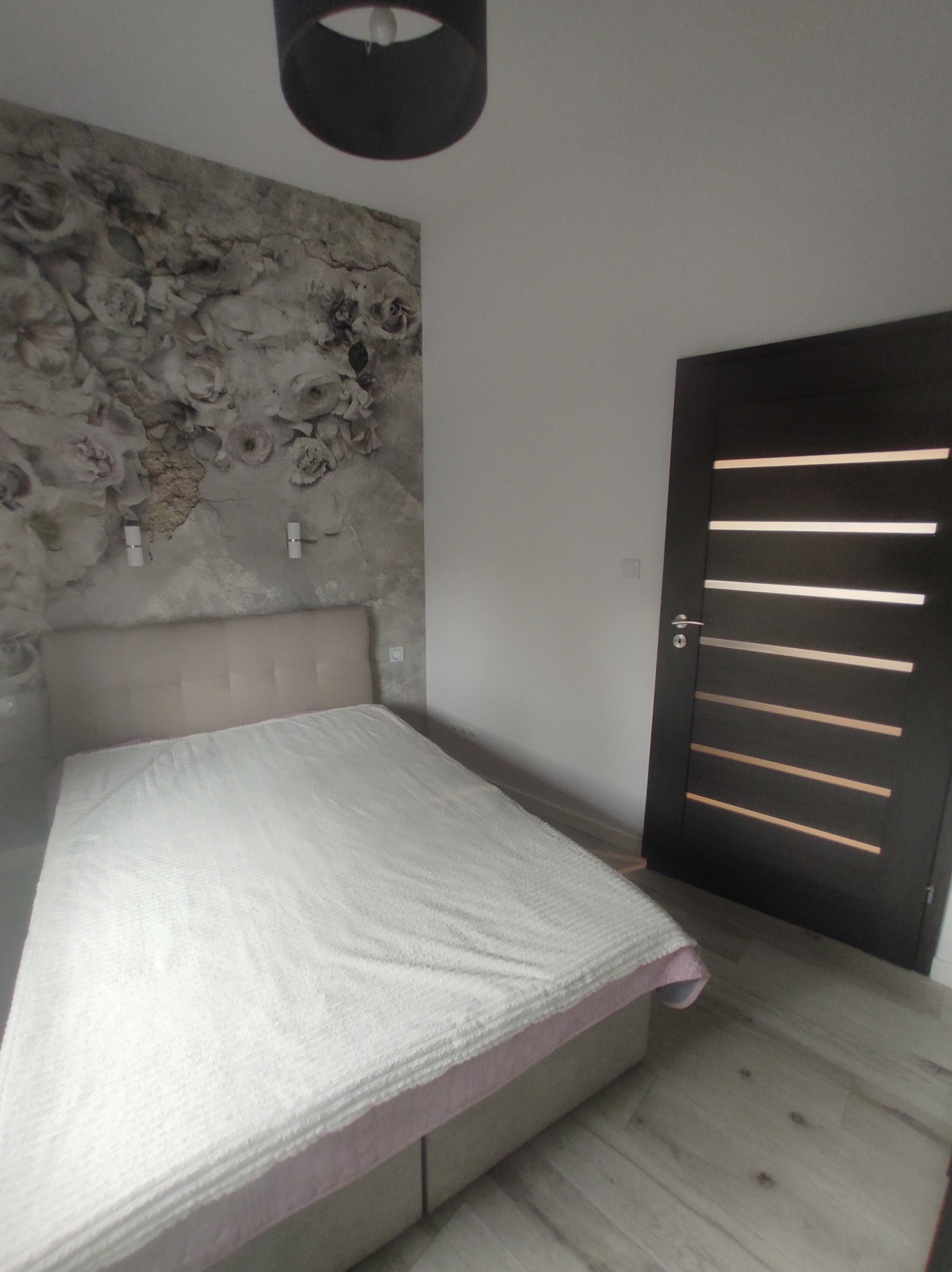 Sypialnia z łóżkiem z beżowym zagłówkiem, białą pościelą z różowym wykończeniem, fototapetą z motywem kwiatowym i ciemnymi drzwiami z poziomymi przeszkleniami.