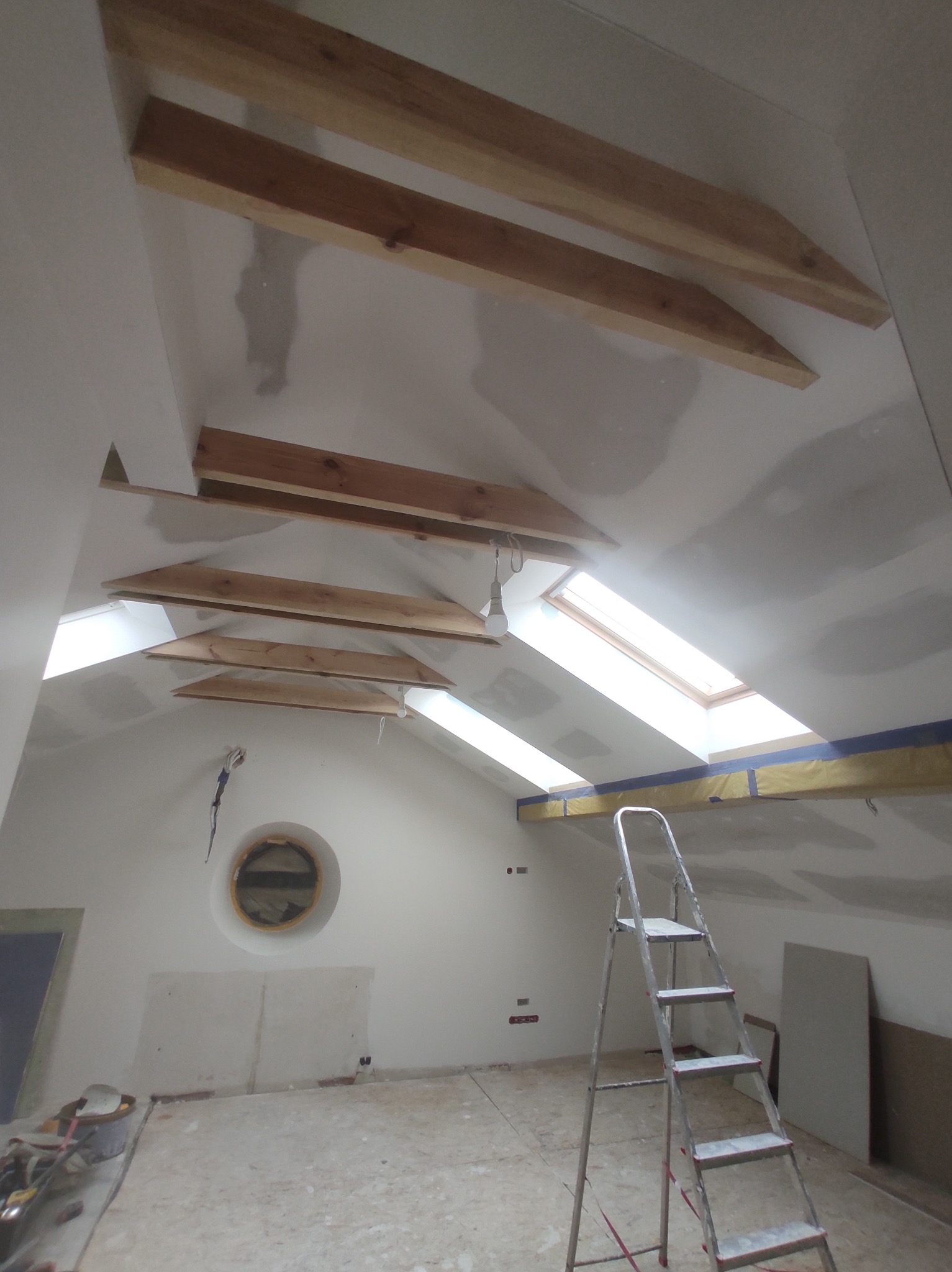 Poddasze w trakcie prac wykończeniowych: widoczne drewniane belki stropowe, okna dachowe, ściany w trakcie szpachlowania, aluminiowa drabina i okrągłe okno.