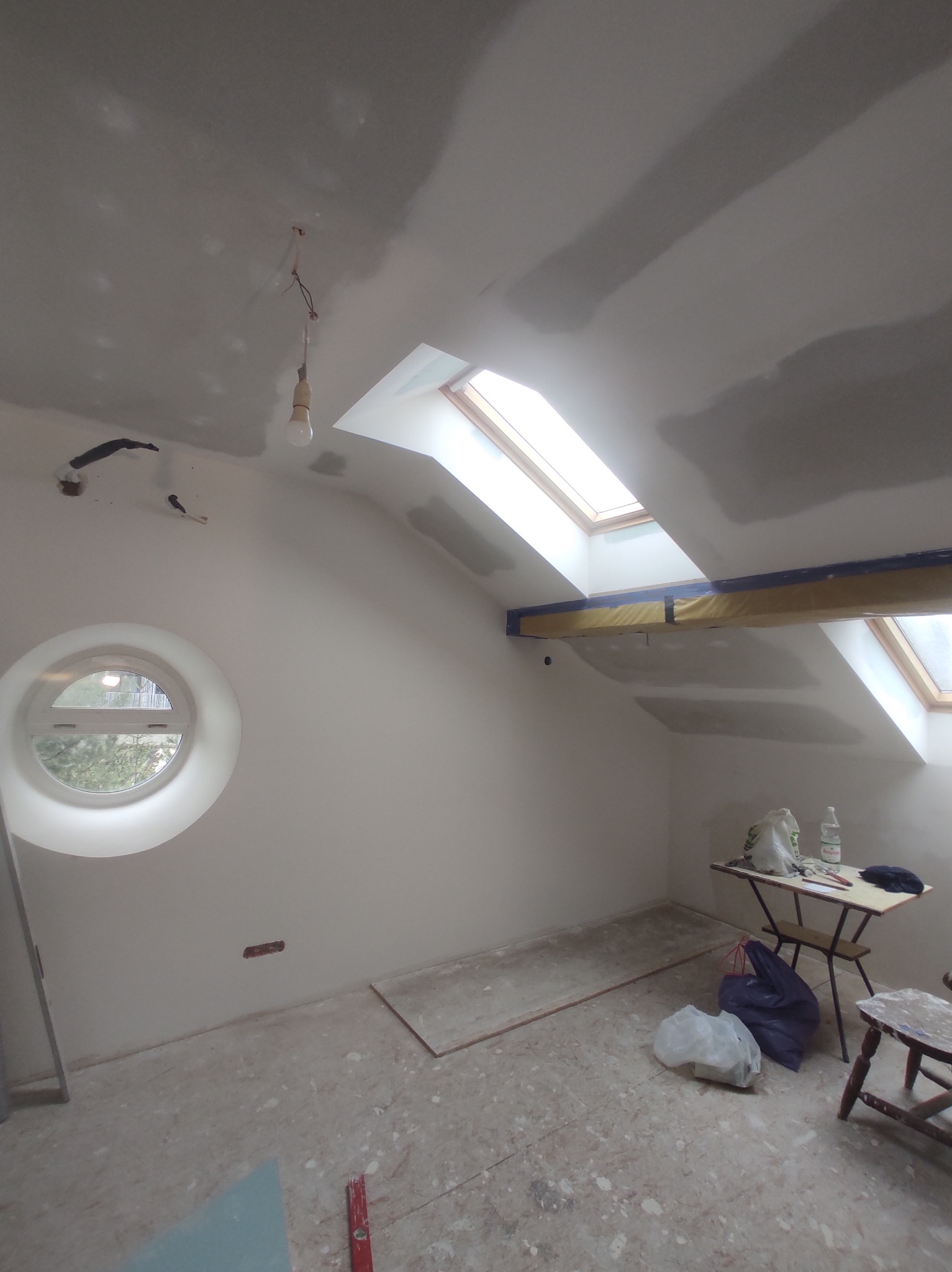 Poddasze w trakcie remontu: ściany po szpachlowaniu, okrągłe okno, widoczne ślady po pracach na podłodze, stolik z narzędziami i materiałami malarskimi.